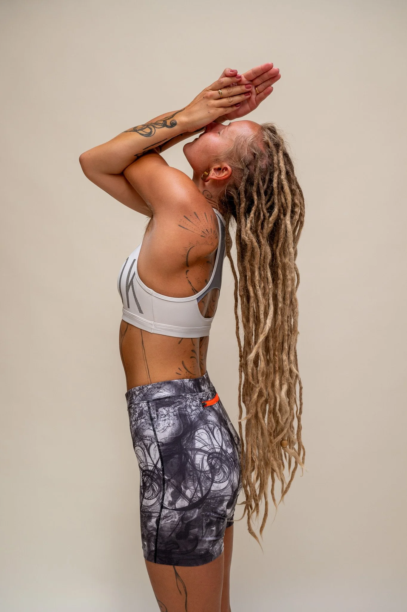 En kvinde med dreadlocks, tatoveringer, og træningstøj, der står mod en neutral væg og laver en yoga-øvelse.