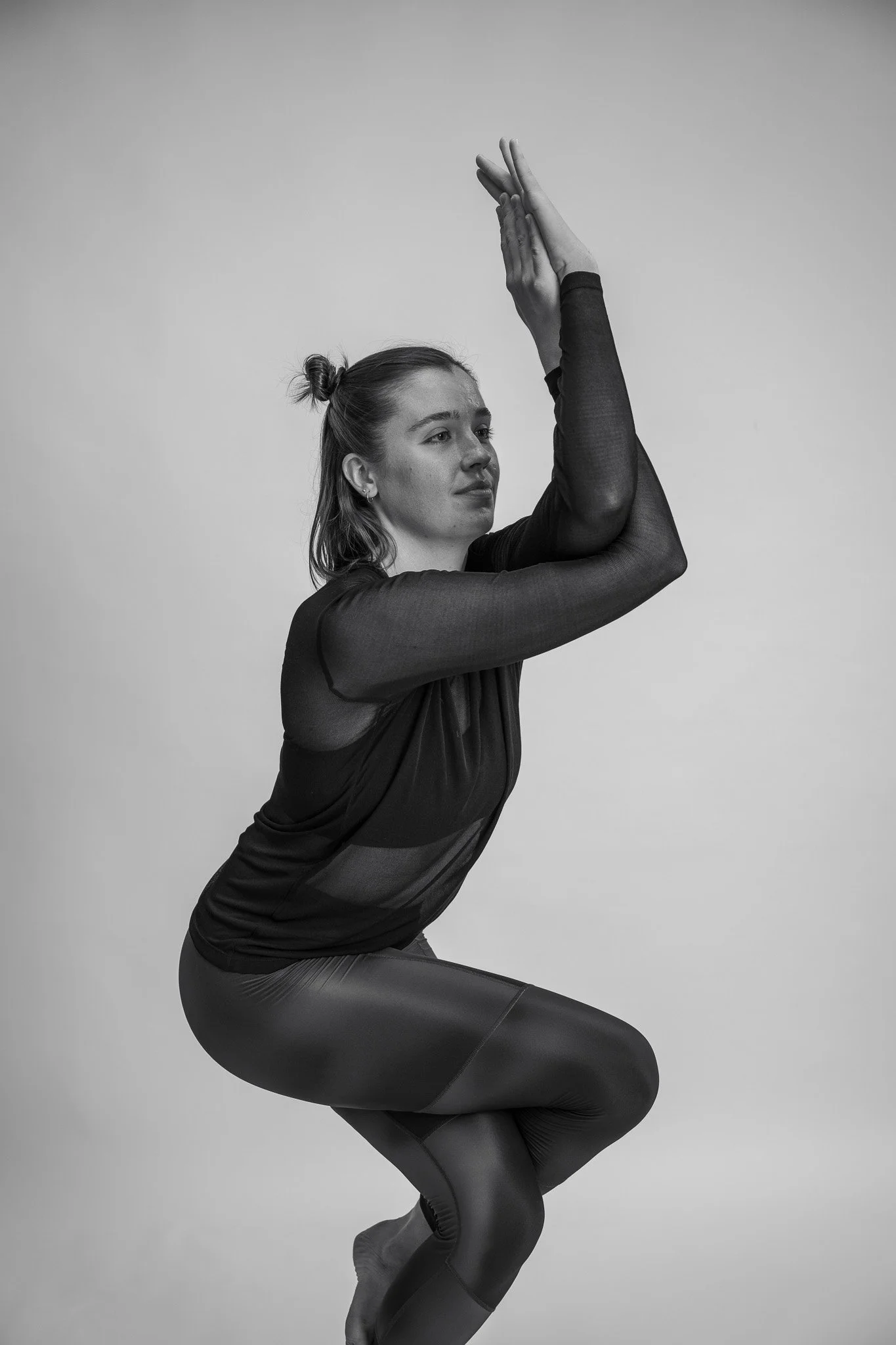 Sort-hvid billede af en kvinde i træningspose, der sidder i en squat holdning med hænder samlet over hovedet.