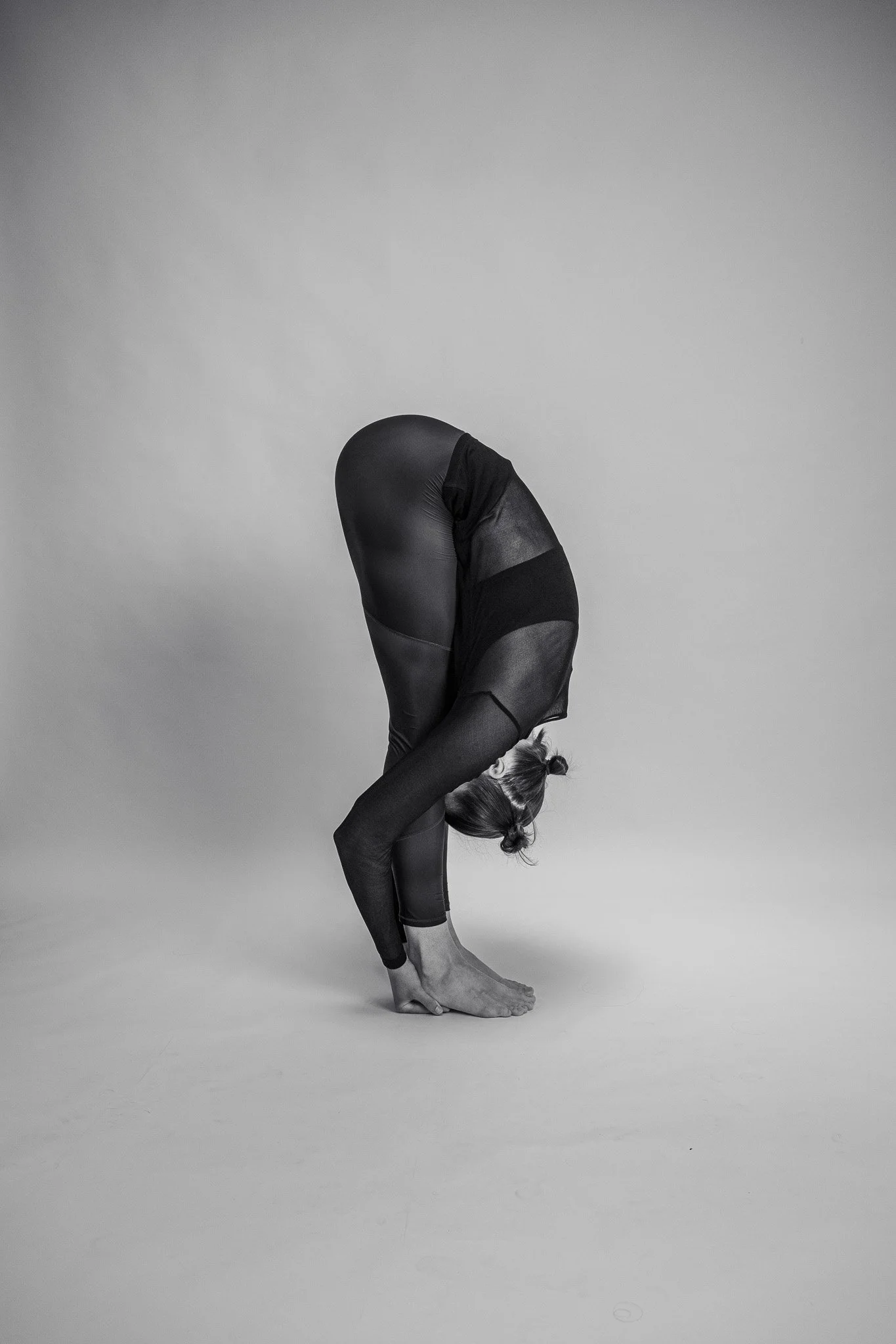 Person i sort tøj, der laver en foroverbøjning med ansigtet mod gulvet, i en yoga- eller strækposition, på en neutral baggrund.