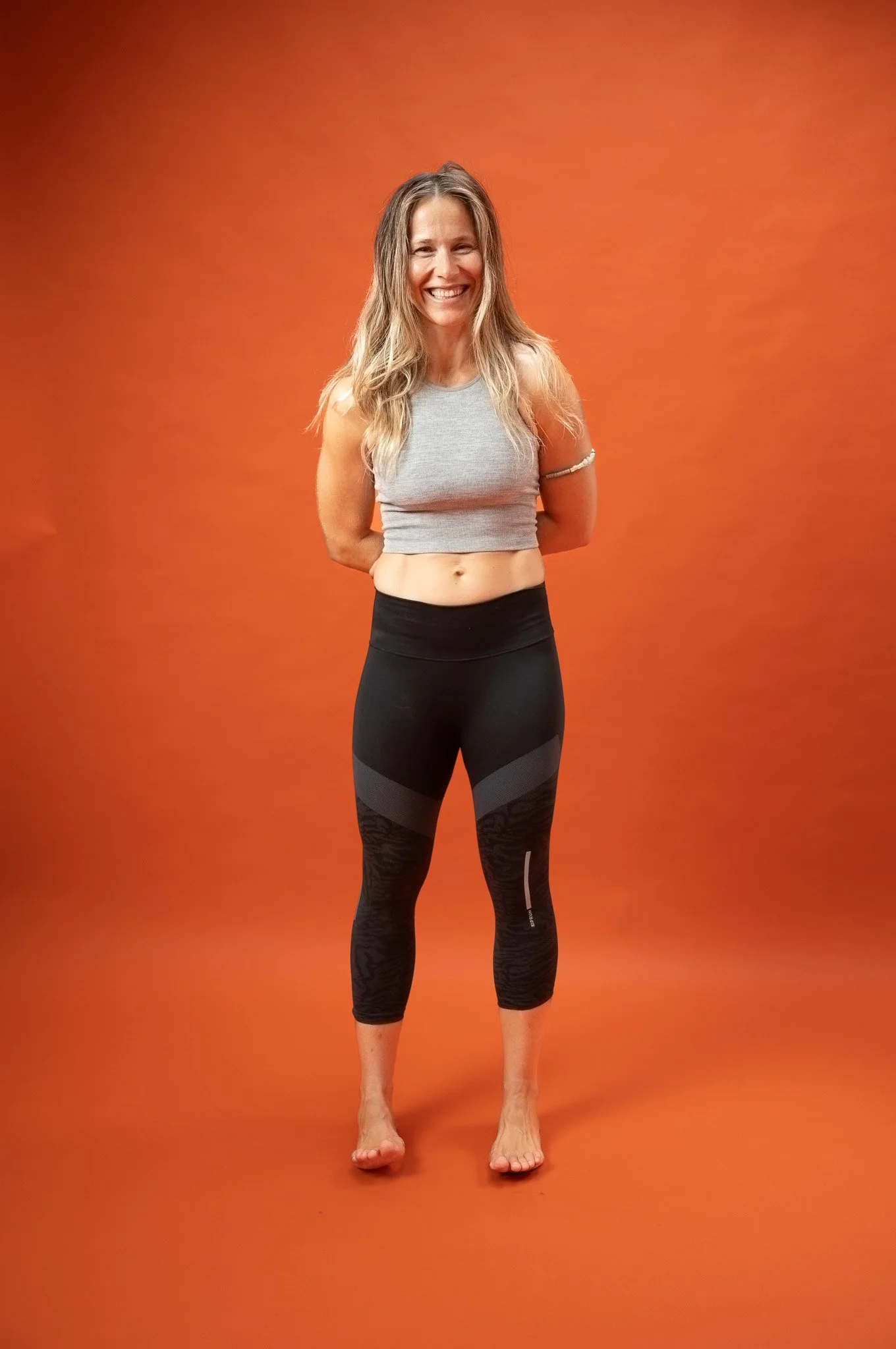 En smilende kvinde står på en orange baggrund iført sportstøj, bestående af en grå top og sorte leggings.