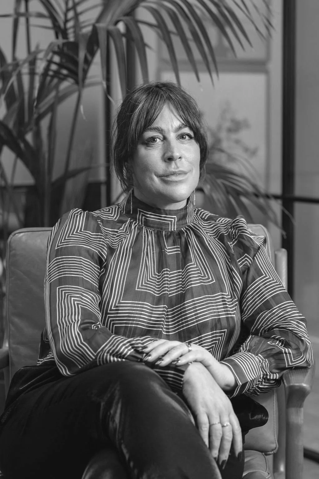 Black and white photo of a woman seated in an indoor setting with large plants in the background. She has short dark hair and is wearing a patterned high-neck blouse, sitting with her hands resting on her lap, looking at the camera.
