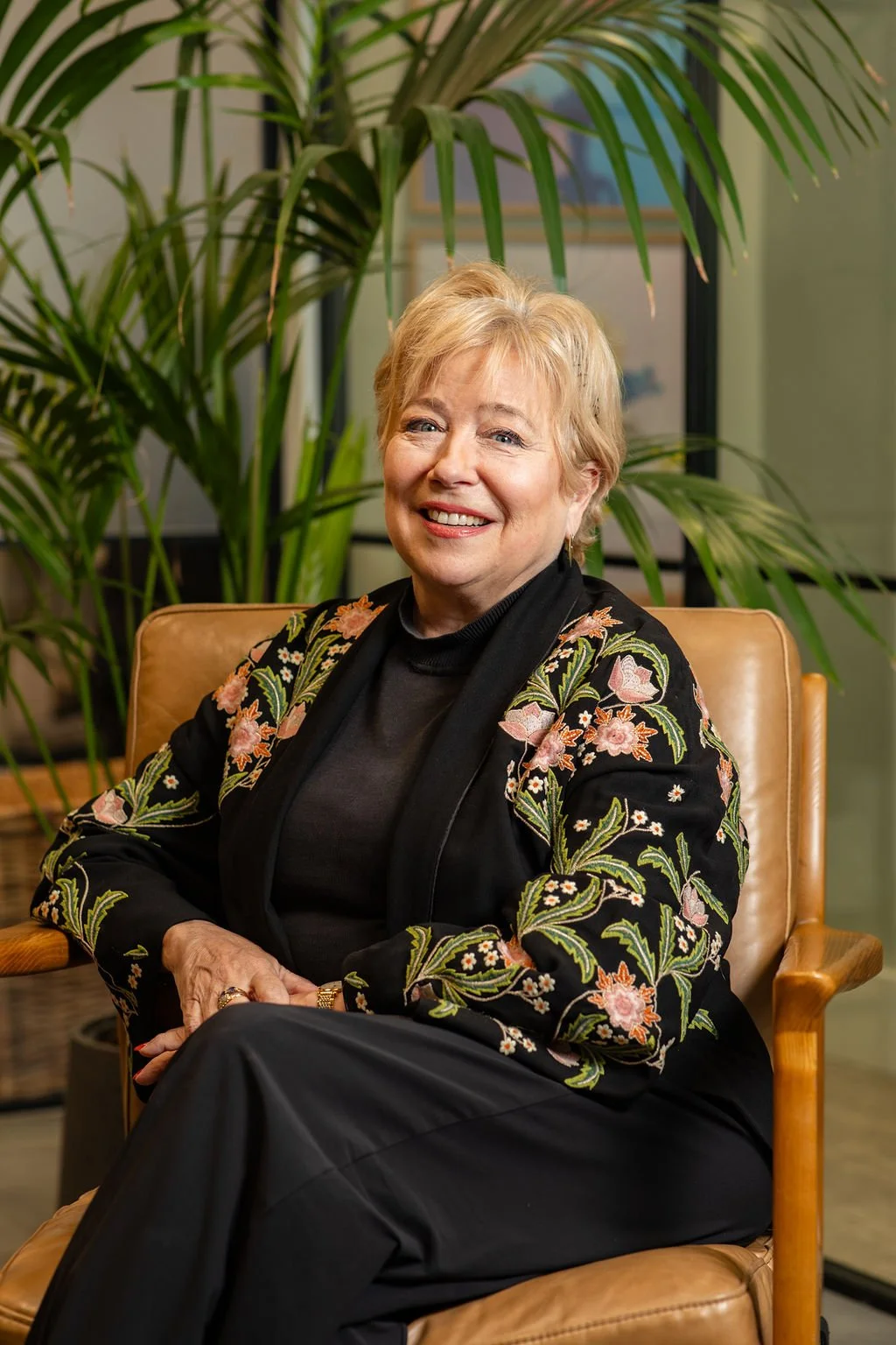 A smiling older woman with short blonde hair, wearing a black floral embroidered blazer and black top, sitting in a tan leather chair in front of green plants and framed artworks.