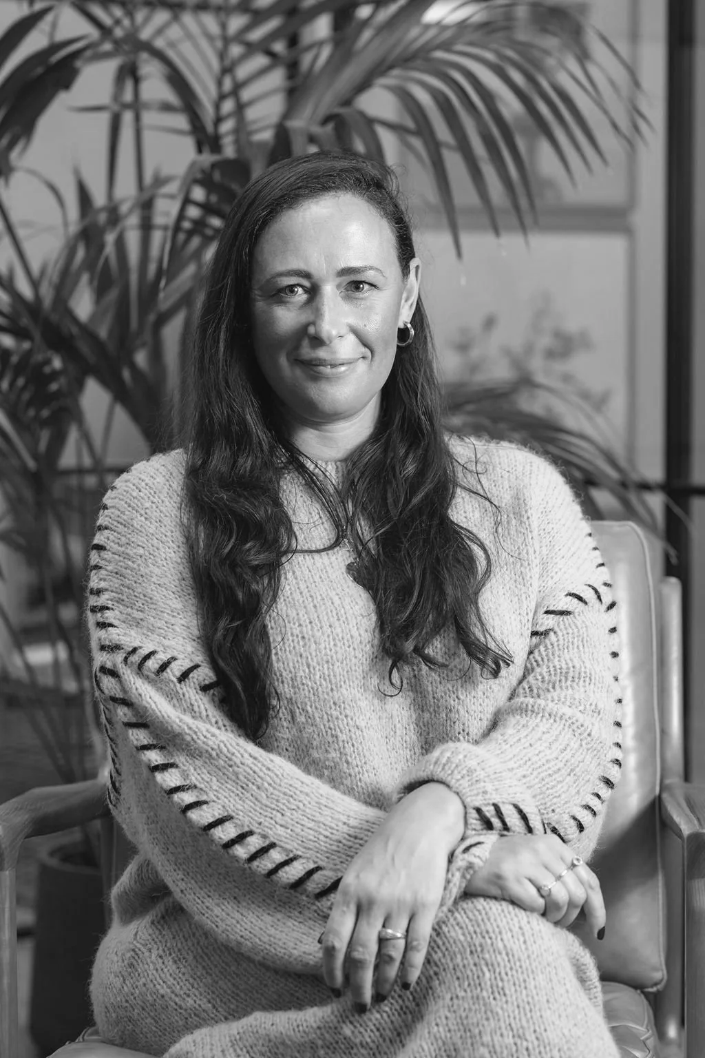 Black and white photo of a woman with long wavy dark hair and hoop earrings, sitting with her arms crossed, wearing a cozy knit sweater, indoors with large leafy plants in the background.