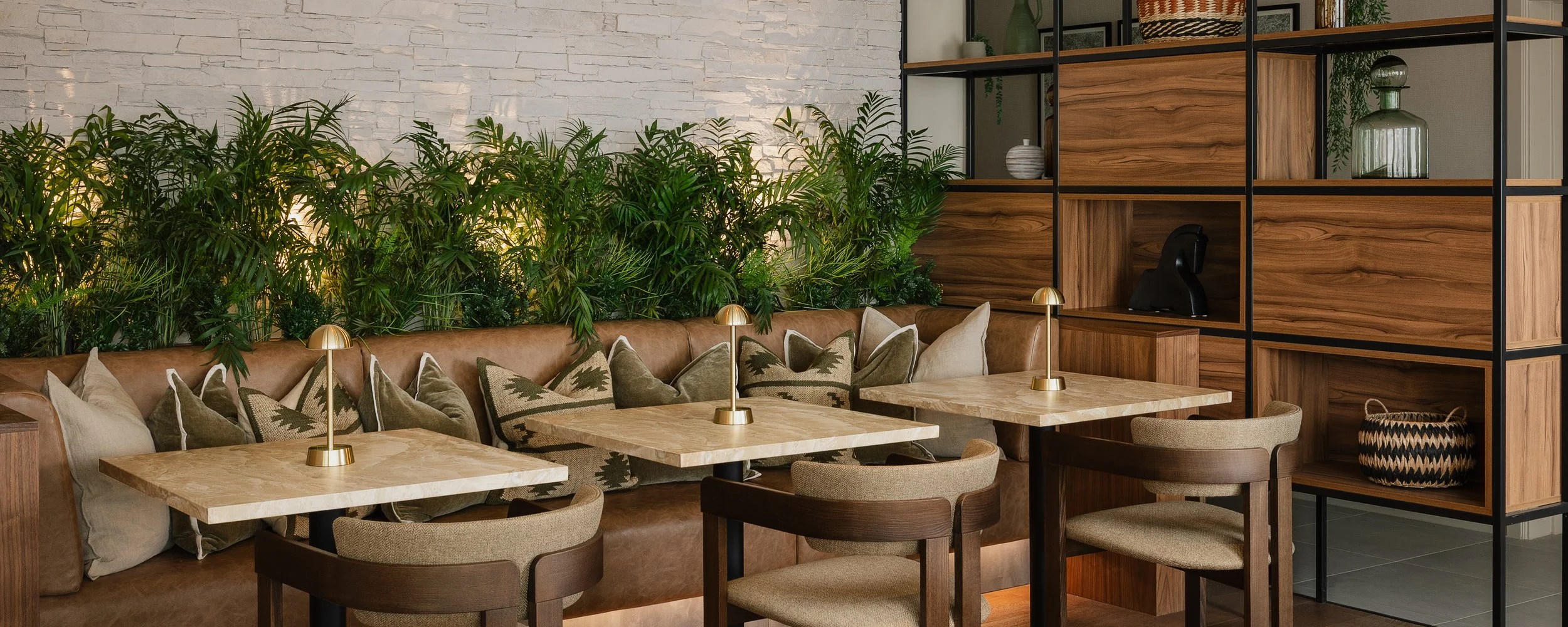 Interior of a stylish cafe with a brown leather booth, decorative pillows, wooden tables with small gold lamps, beige chairs, and a wooden shelving unit with decor, greenery, and glass jars.
