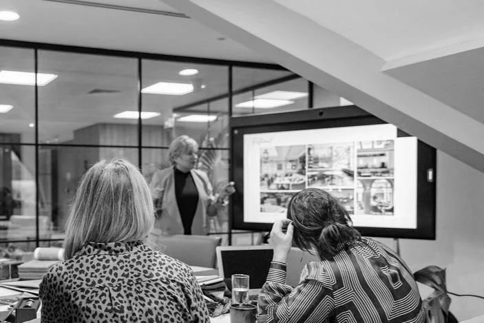Two women sitting at a table in a conference room, looking at a presentation on a large screen, with a woman standing near the screen.