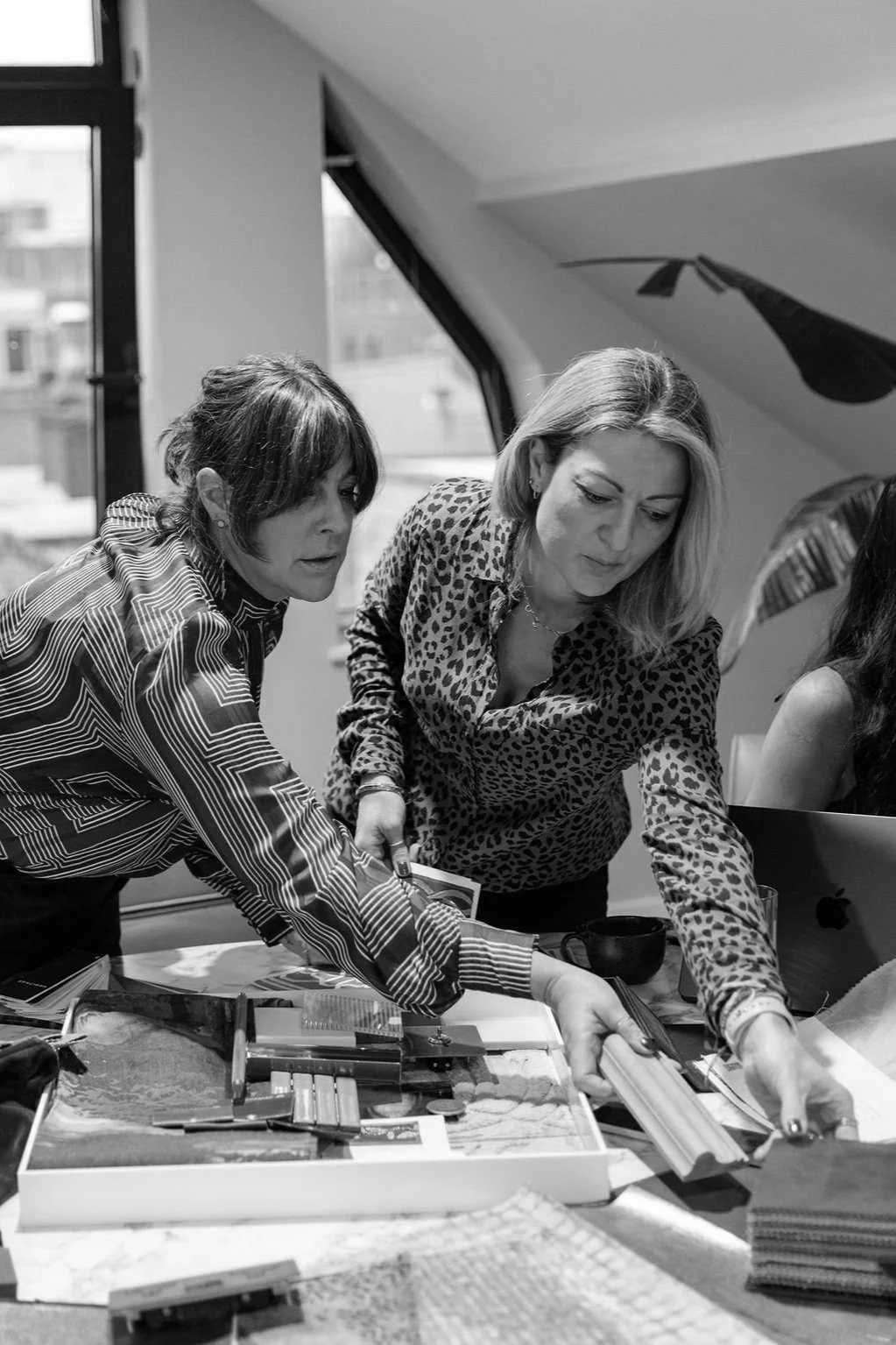 Two women with patterned shirts organizing fabric swatches and samples on a table in an office setting.