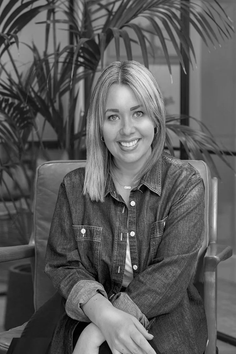 Black and white photo of a smiling woman with shoulder-length hair, seated in a chair with a large plant in the background.