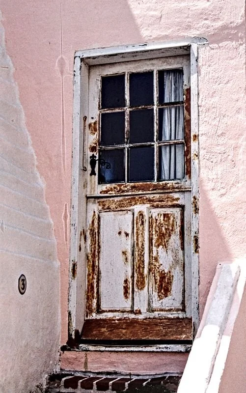 Old, rusty white wooden door with glass panes, set in a pink stucco wall, with a small step at the bottom.