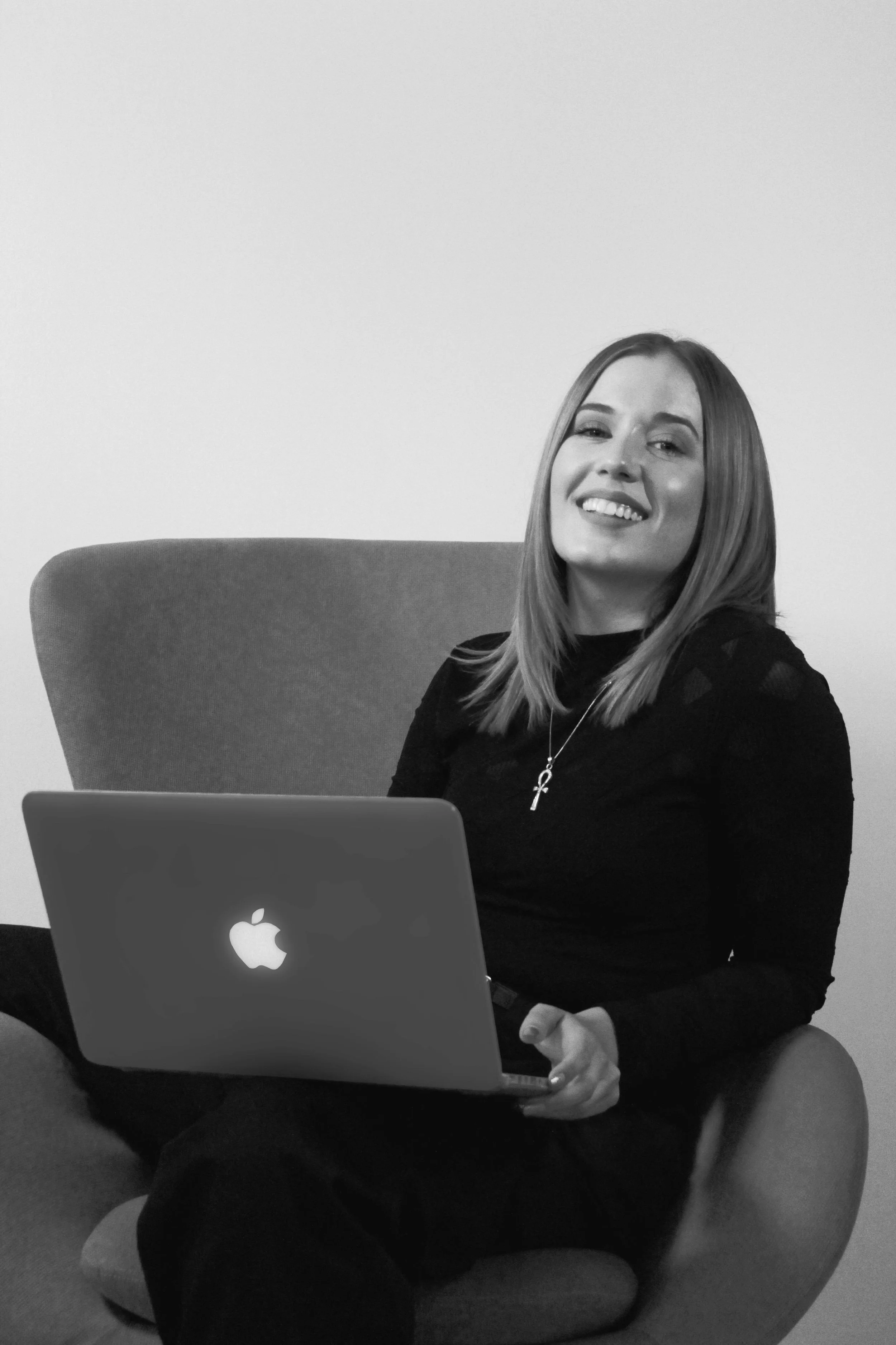 Een lachende vrouw met lang haar zit op een comfortabele stoel, houdt een laptop met een Apple-logo op schoot en kijkt naar de camera.
