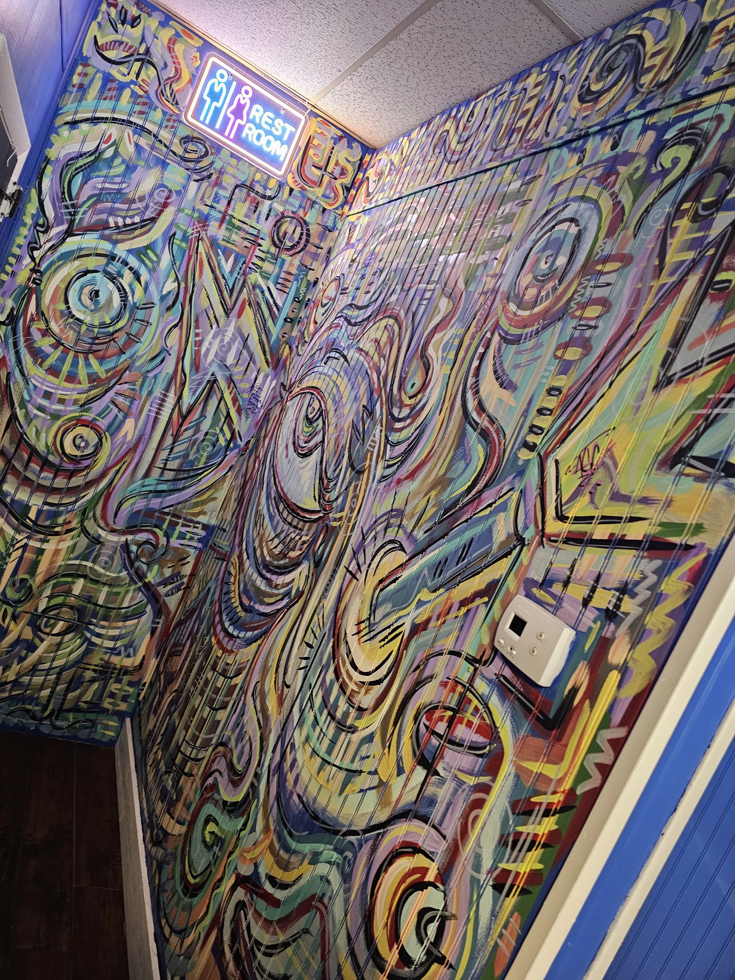 Interior wall with colorful abstract graffiti painting, finished with black lines and various bright colors. There is an illuminated rest room sign at the top corner.