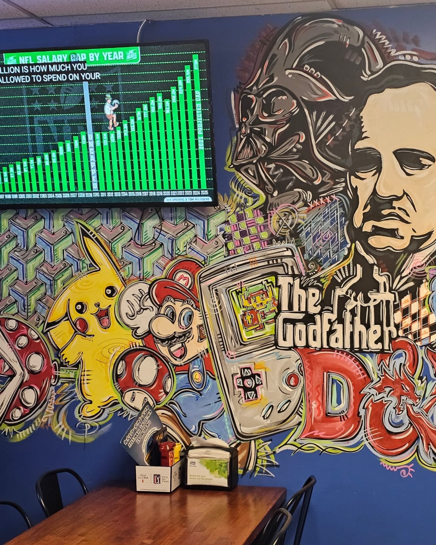 Colorful graffiti mural with cartoon characters, a large face, and a sign reading 'The Godfather' on a wall behind a wooden table and chairs.