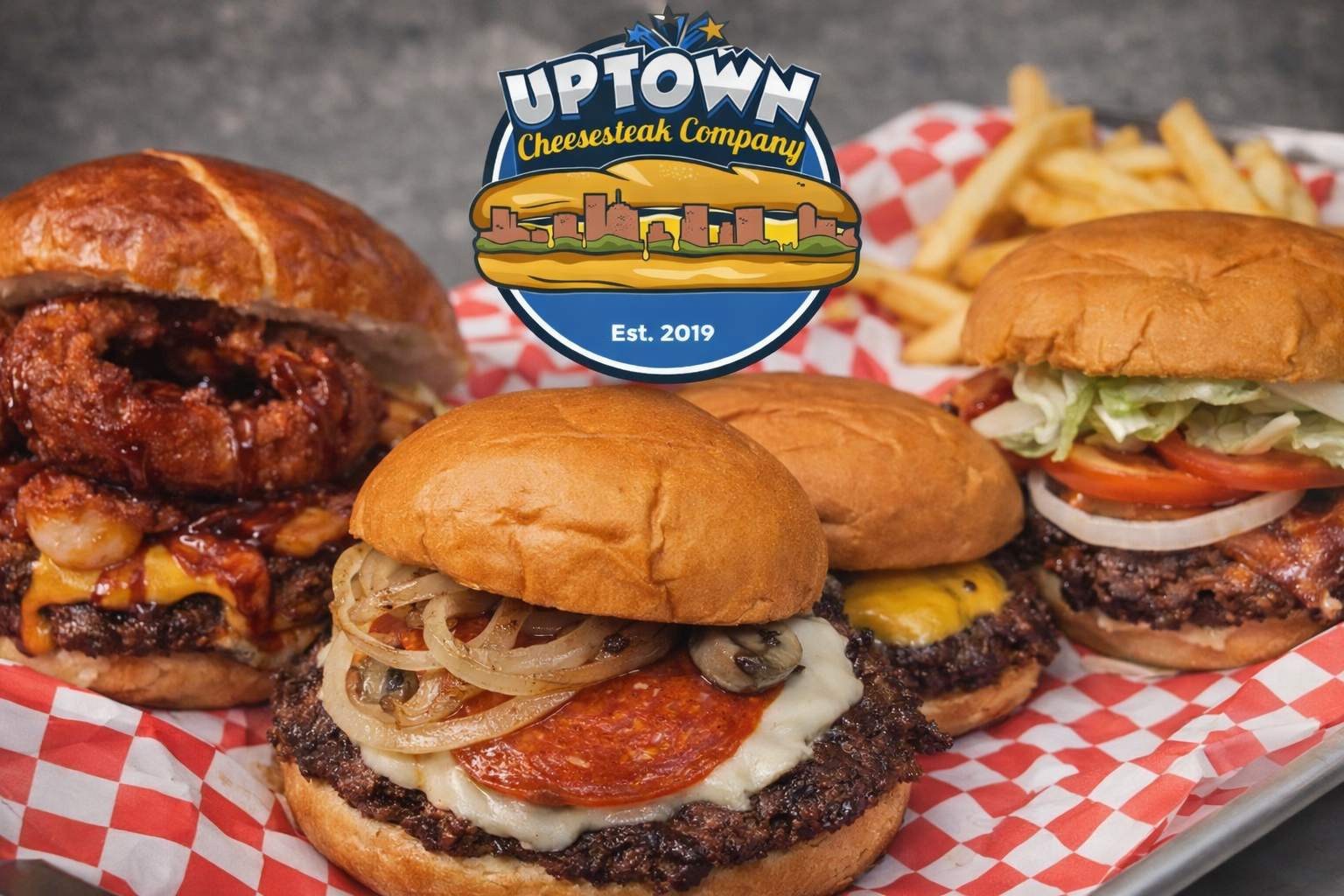 Variety of cheeseburgers with toppings on a red and white checkered paper with a tray of French fries in the background, and a logo of uptown cheesesteak company with the text understated 2019.