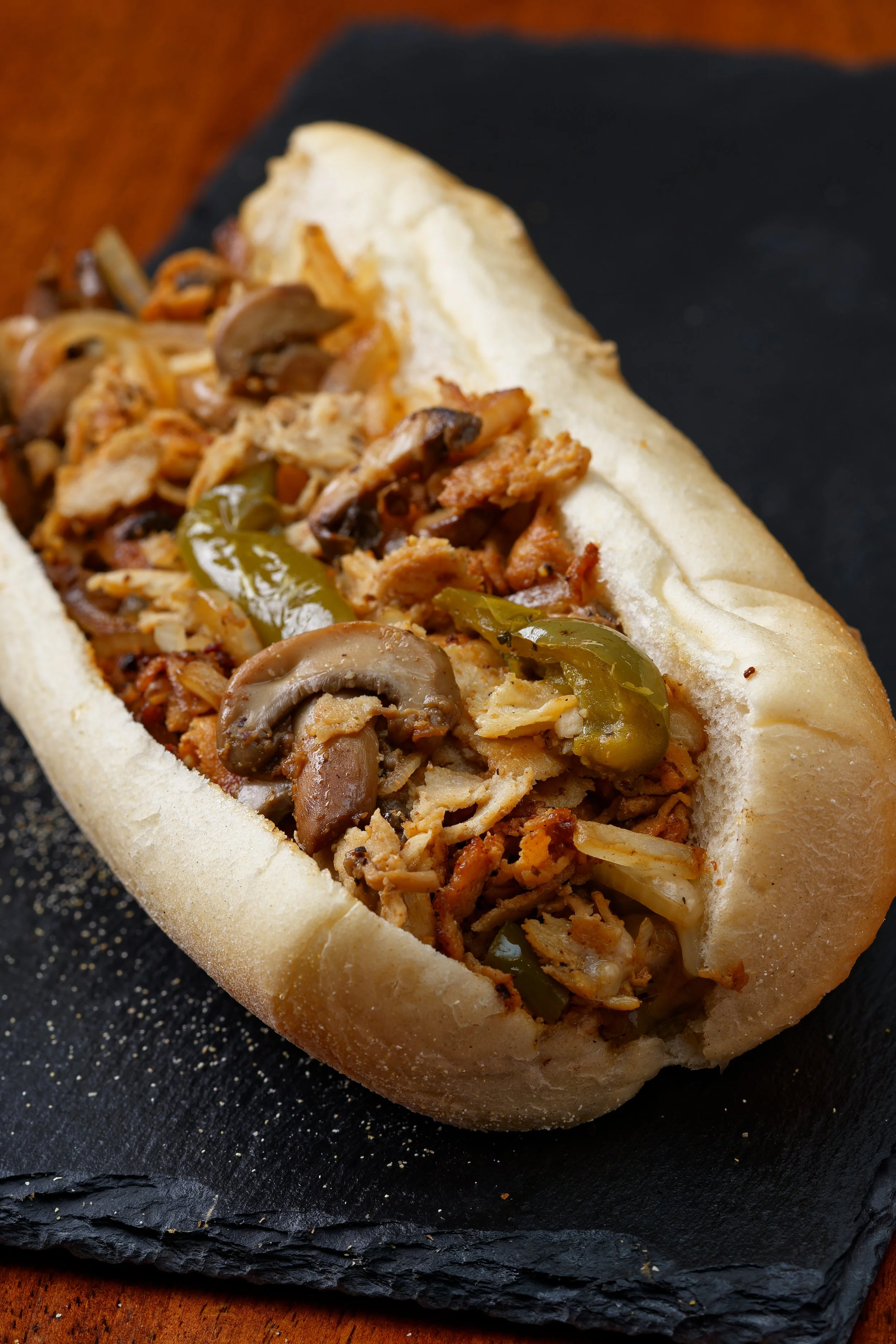 Hot sausage and mushroom hoagie sandwich on a white hoagie bun with sautéed green peppers and onions, served on a black slate board.
