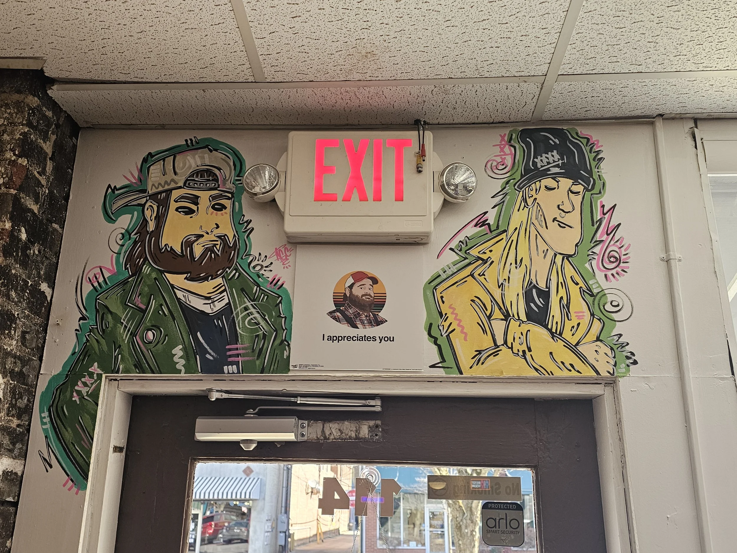 Graffiti artwork of two people on the wall near an exit sign and door. The person on the left is a man with long hair and a beard, wearing a green jacket and a backwards cap. The person on the right is a woman with long blond hair, wearing a black ca
