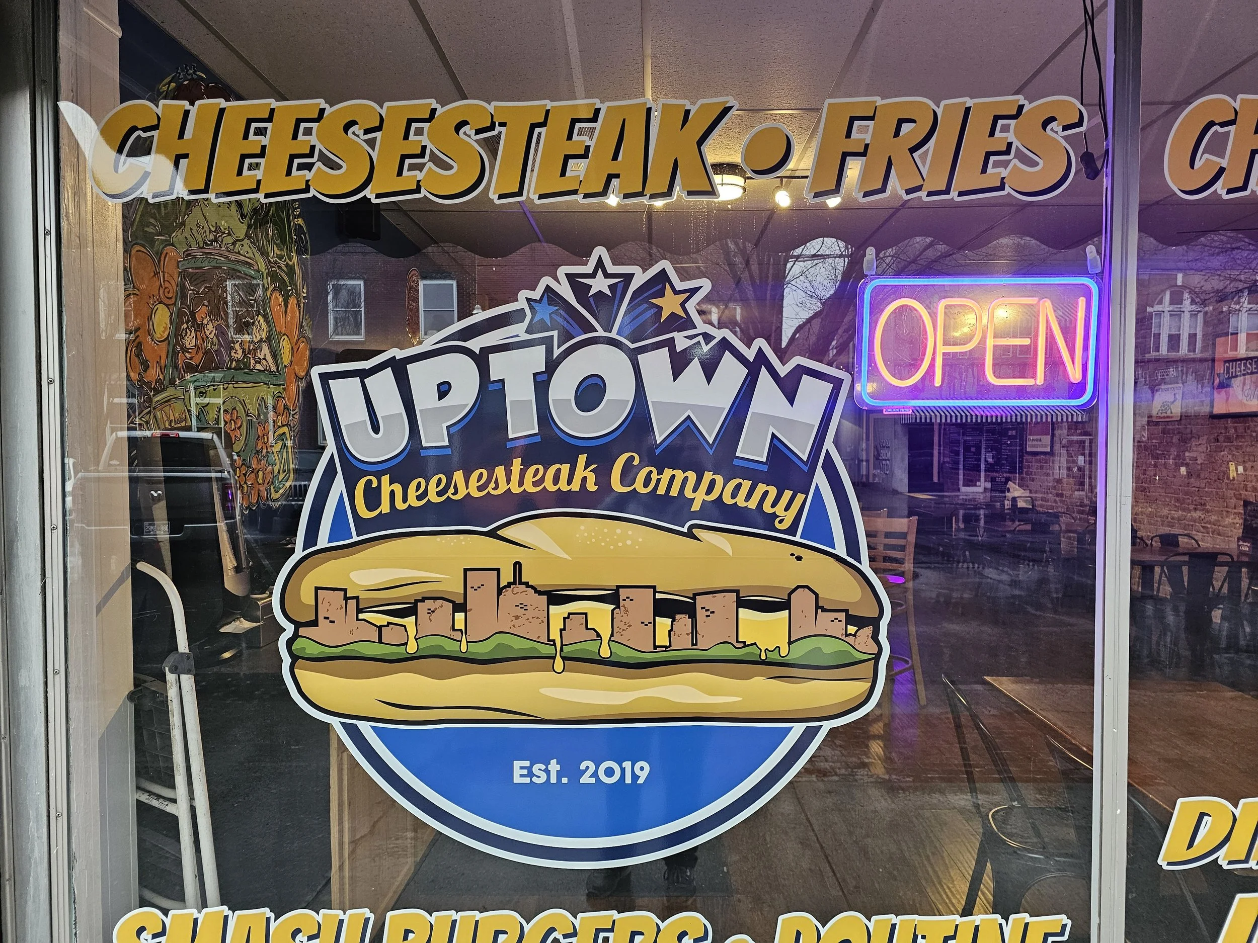 A storefront window advertising cheesesteaks, burgers, and chicken. The sign features a large cheeseburger graphic with city skyline at the bottom and fireworks above. There is a neon "Open" sign, and the smaller text indicates the establishment was established in 2019.