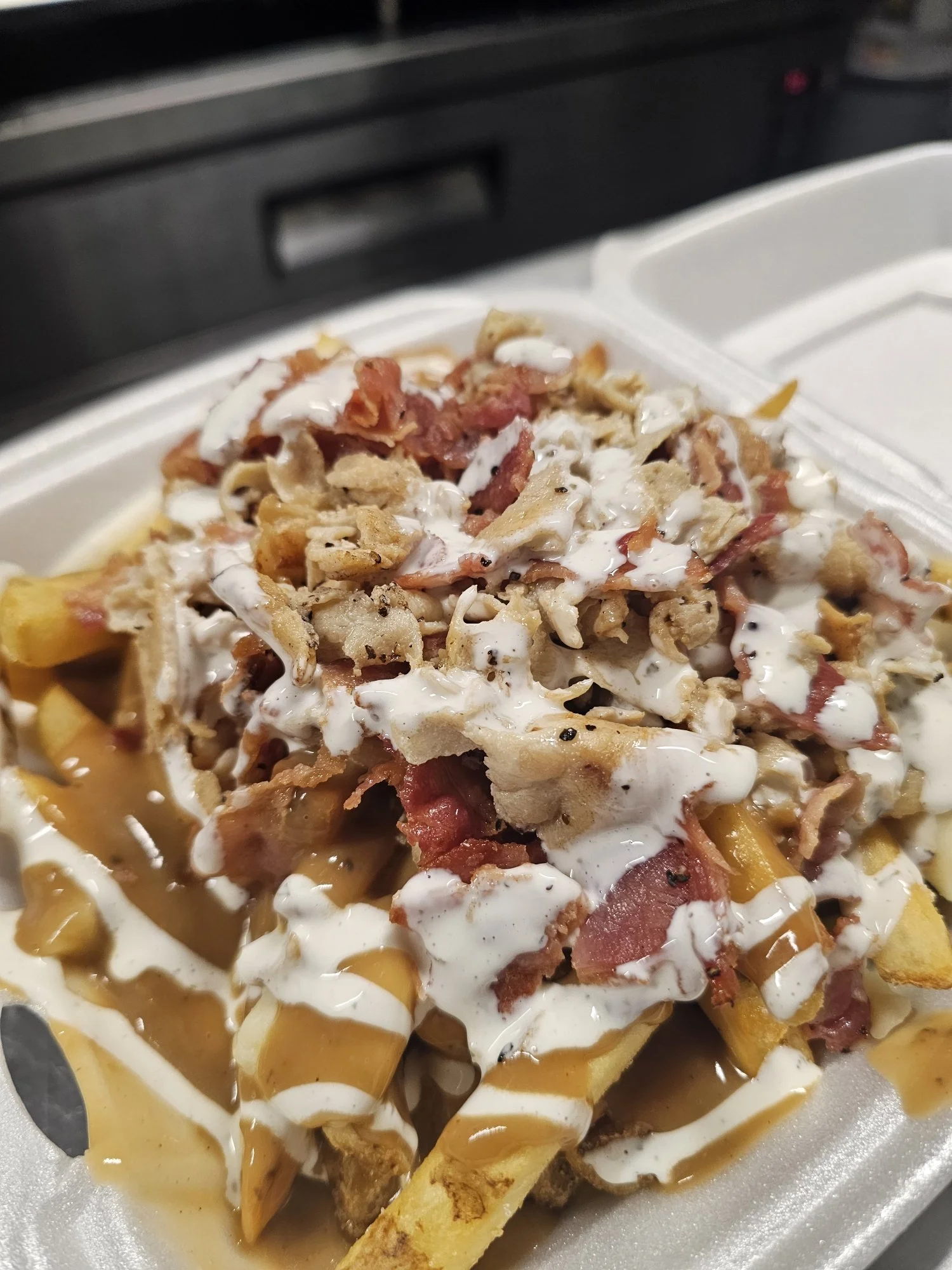 Fries topped with shredded chicken, bacon, cheese, and drizzled with ranch and barbecue sauce in a takeout container.