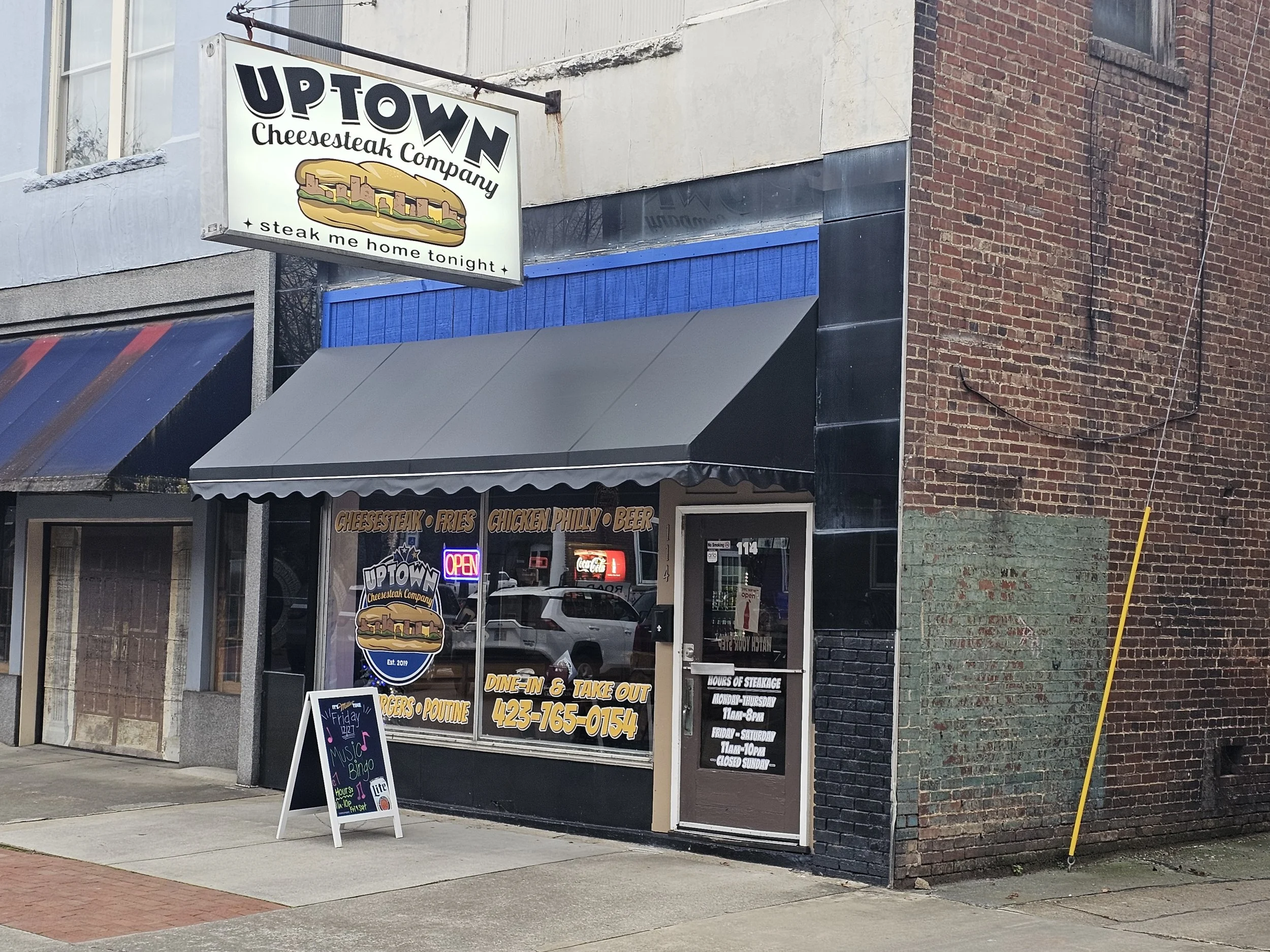 Exterior of a restaurant called UPTOWN Cheesesteak Company with a sign promoting steaks. The storefront features large glass windows, black and blue decorations, and a sidewalk chalkboard with purple and pink music note graphics advertising music bin