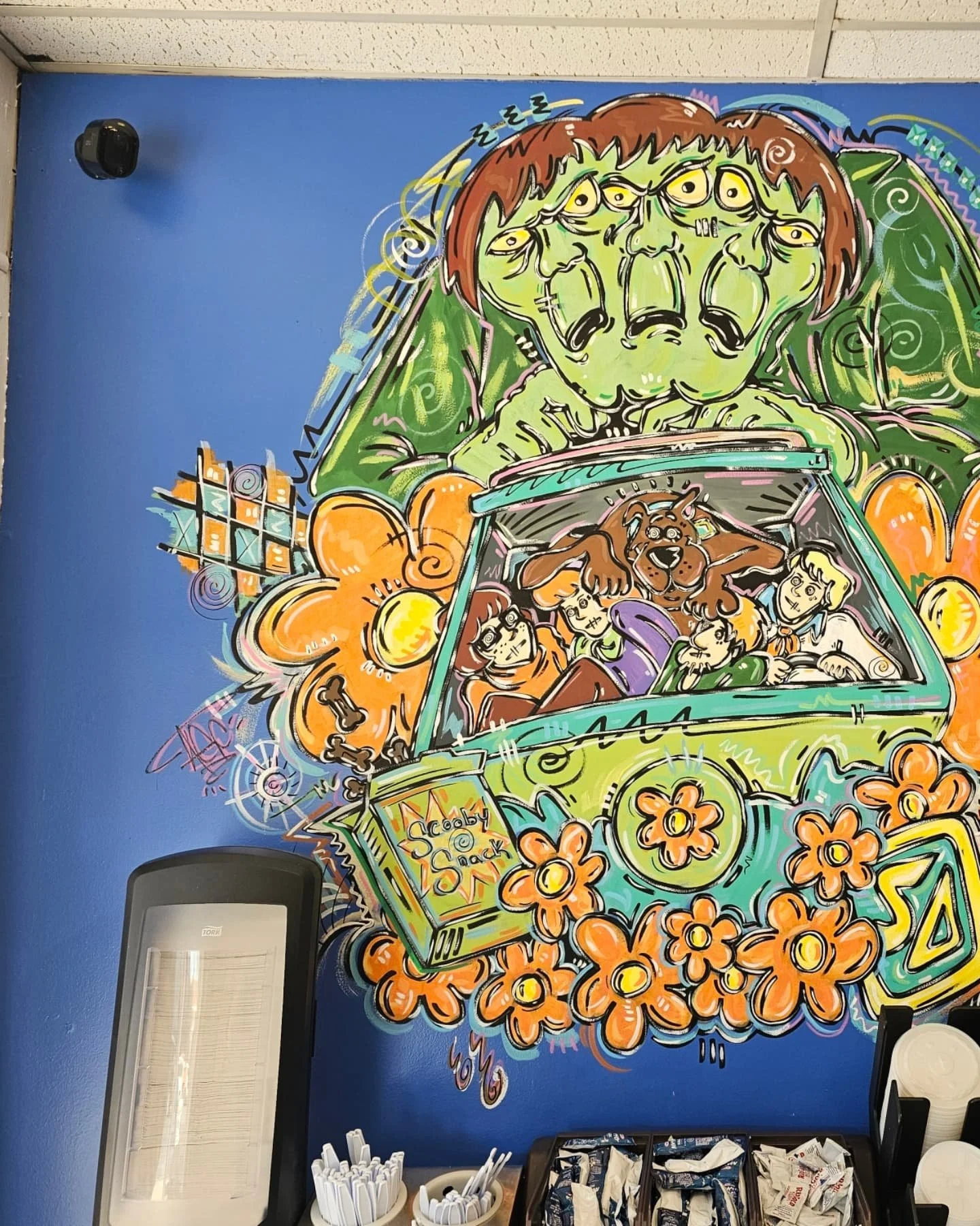 Colorful mural on a blue wall depicting a large green creature with multiple eyes and a wide mouth, in a car with four passengers and a box of snack packets labeled "Scoby Snack" in front.