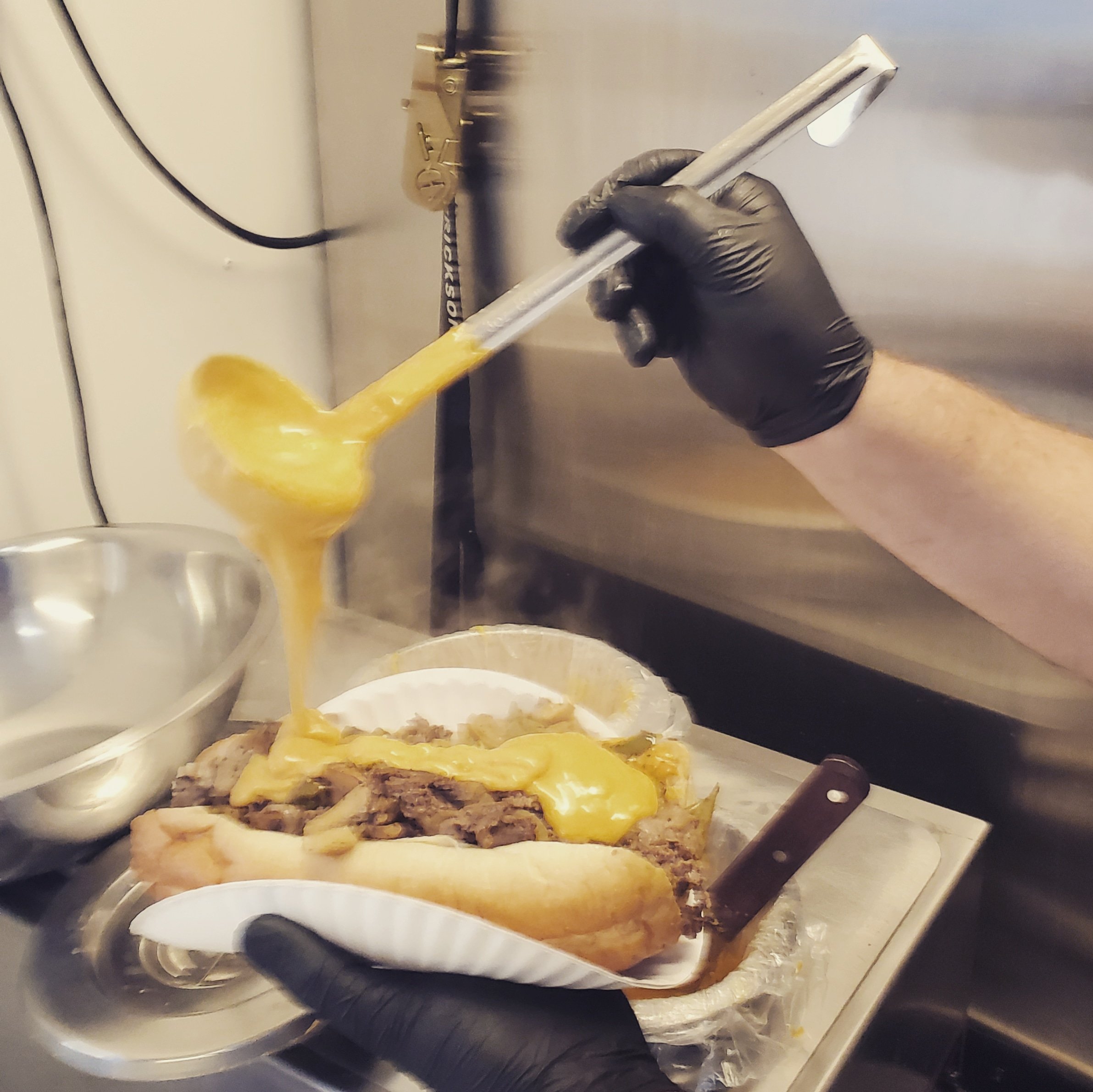 A person wearing black gloves holds a metal spatula and is spreading melted cheese over a freshly baked pizza in an industrial kitchen with stainless steel appliances.