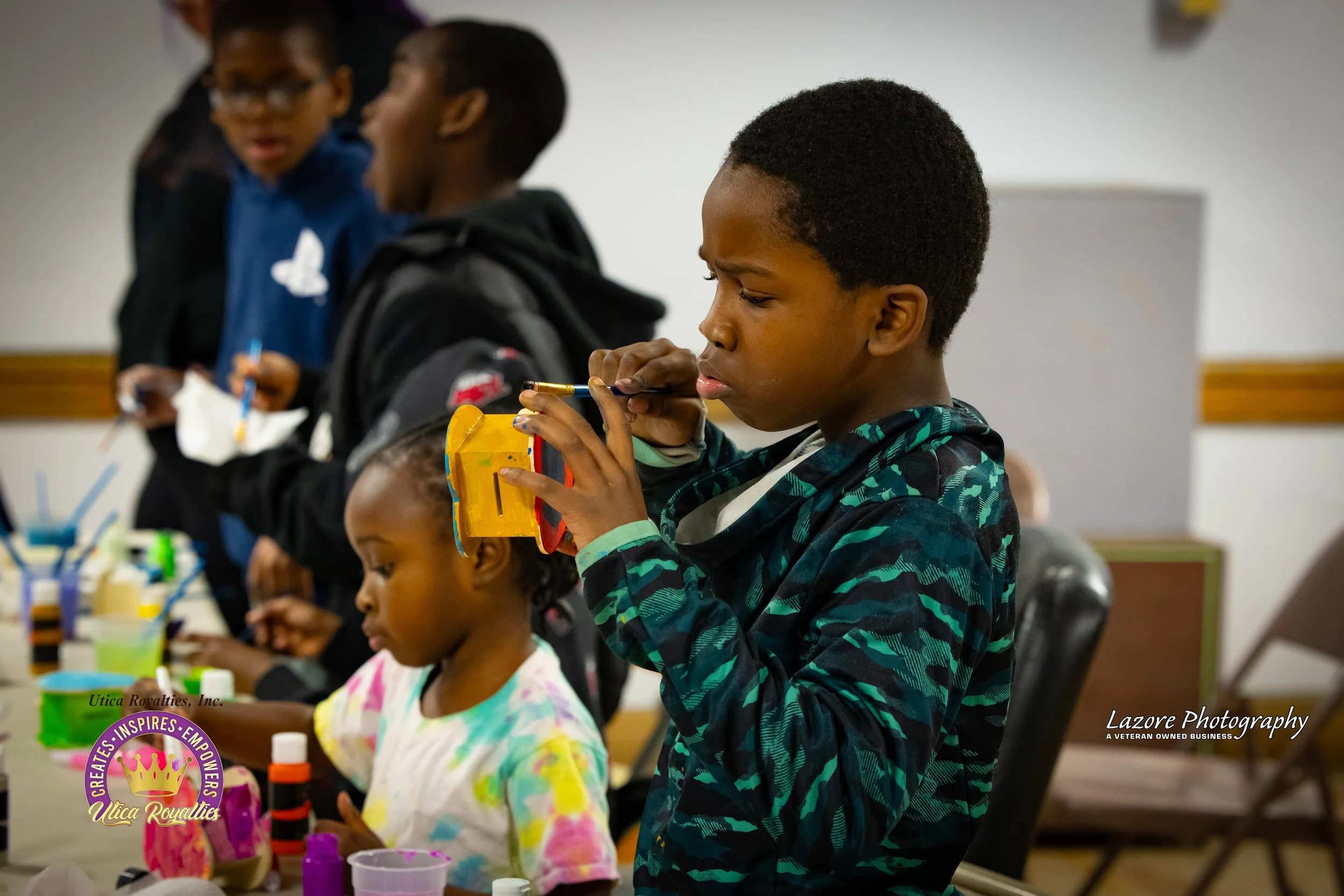Children creating arts and crafts at a table, focusing on a young boy examining a painted object with a paintbrush, surrounded by art supplies and other children working on projects.