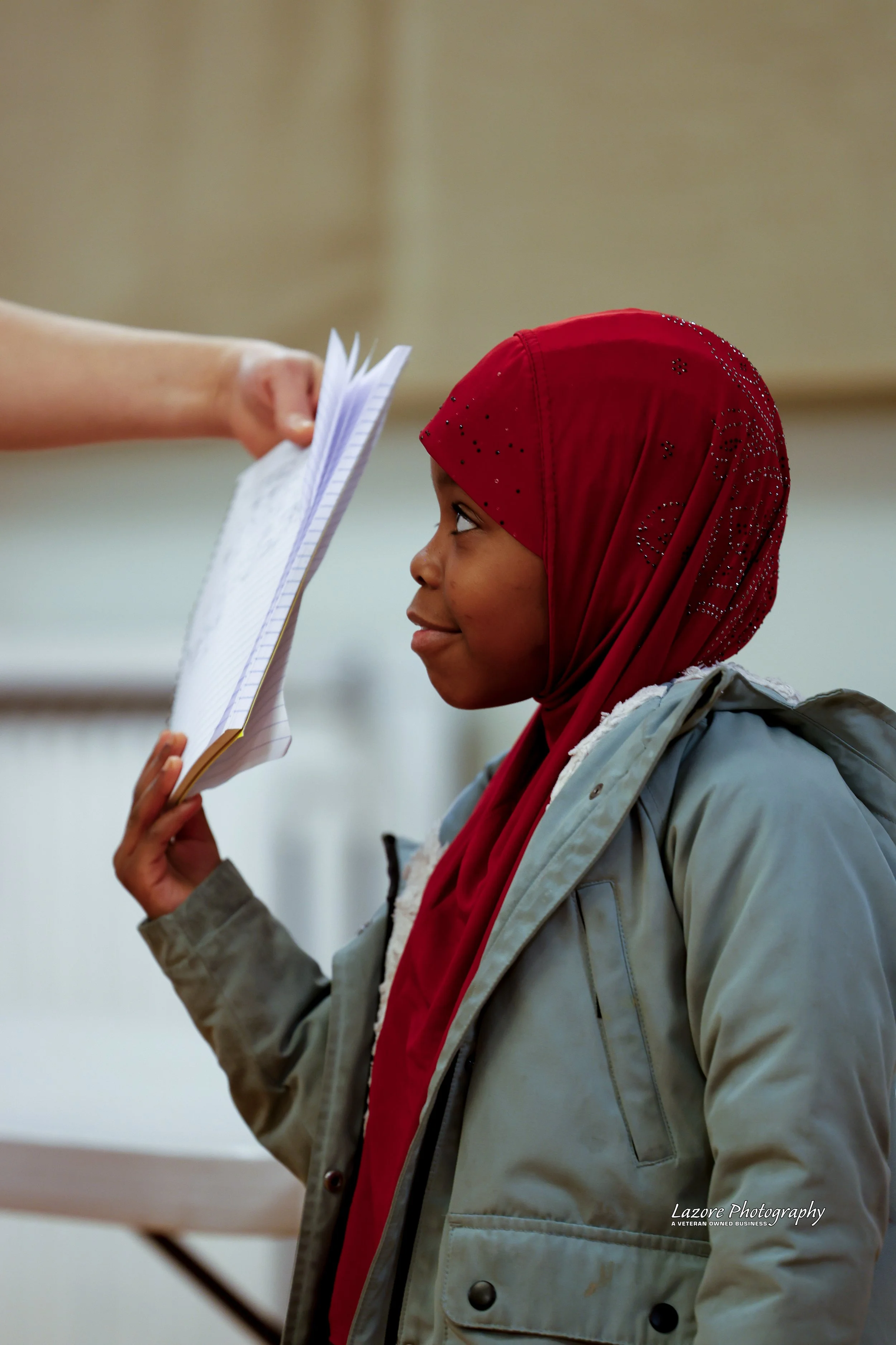A young girl wearing a red hijab and gray jacket is in a classroom, holding a notebook, while someone hands her another notebook or paper. The girl is looking at the person with a slight smile.