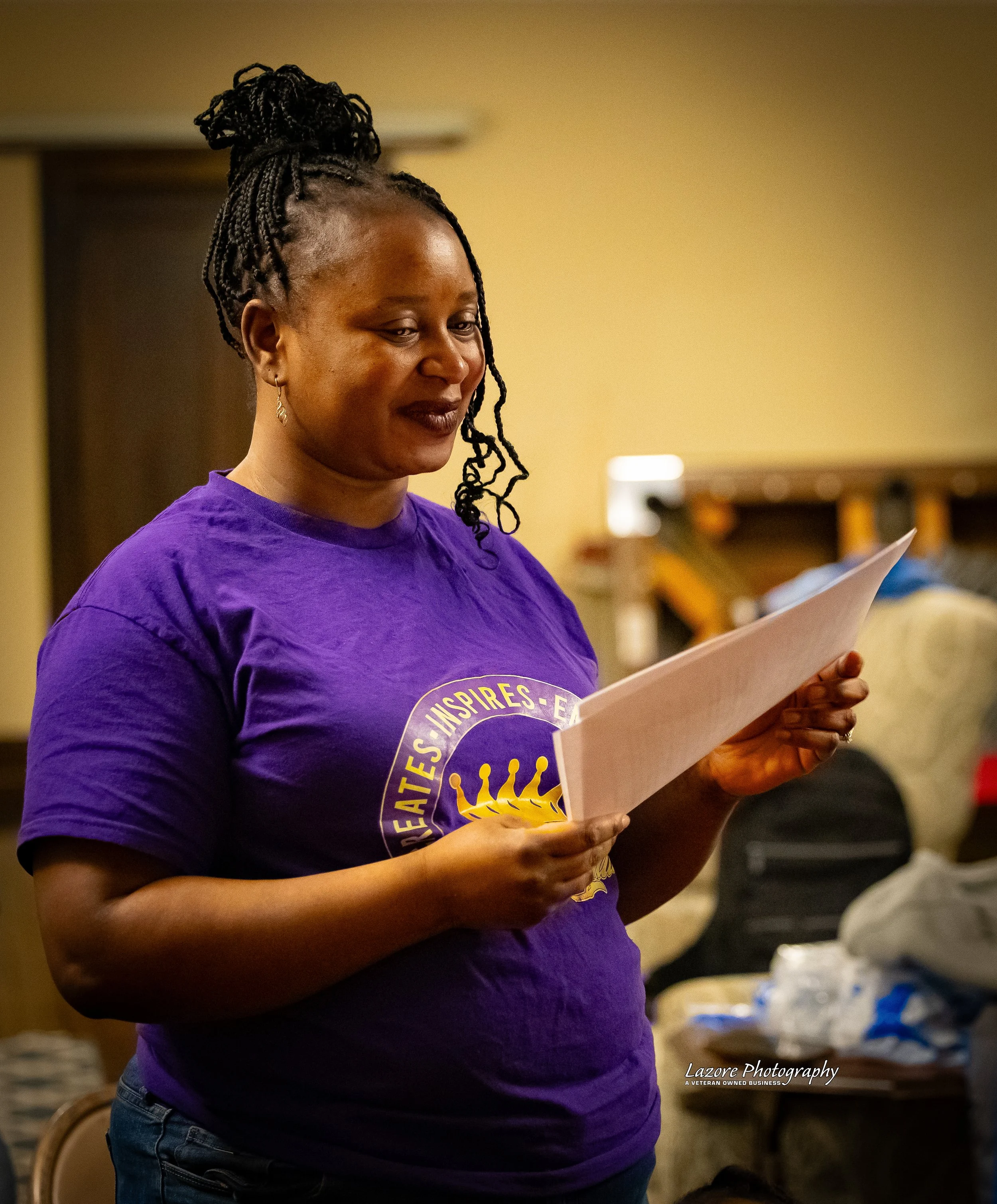 A woman with braided hair and wearing a purple T-shirt with a logo is reading a paper, standing indoors against a blurred background with some items on a table.