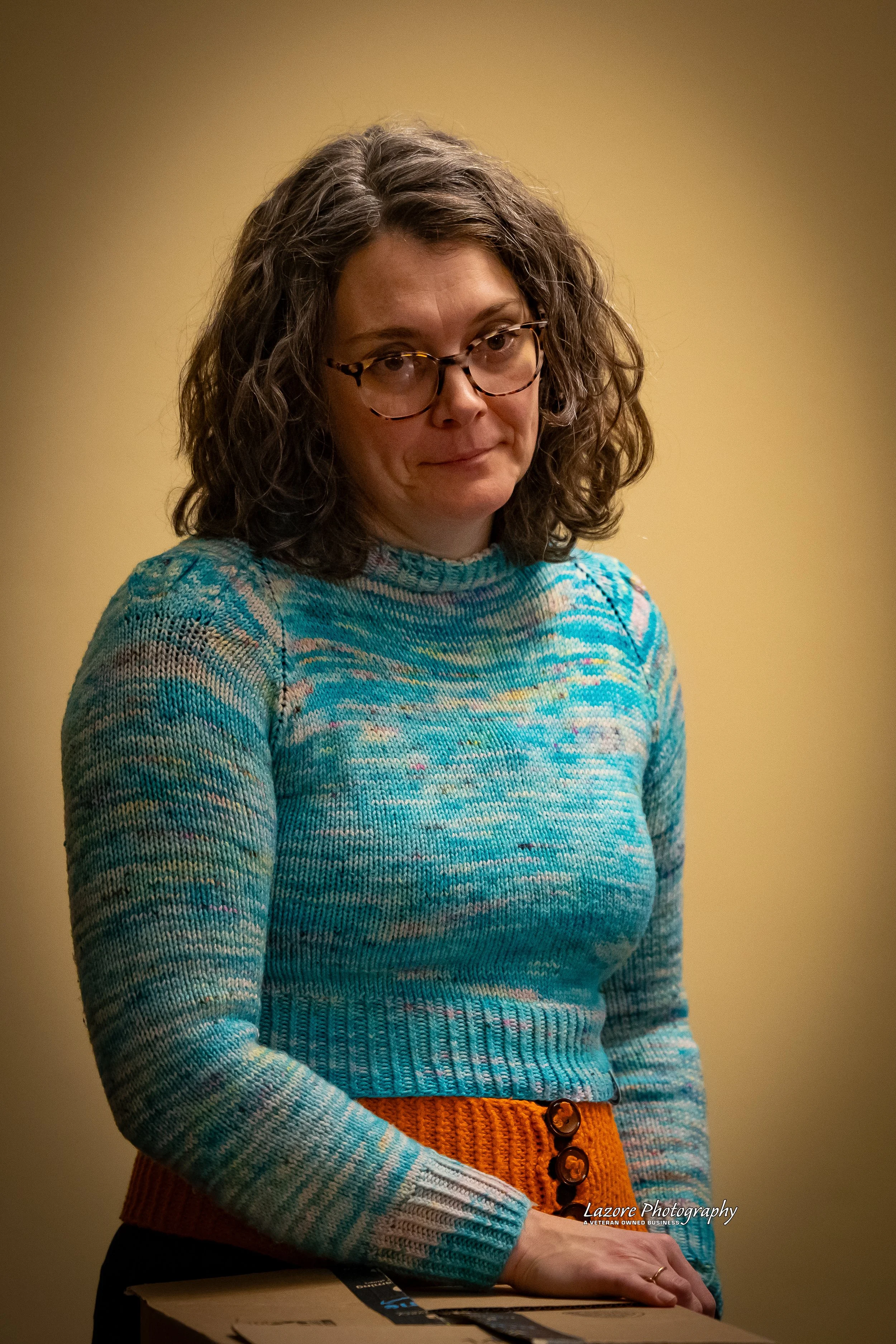 A woman with curly brown hair and glasses, wearing a multicolored blue sweater and orange skirt, standing indoors against a yellow wall.