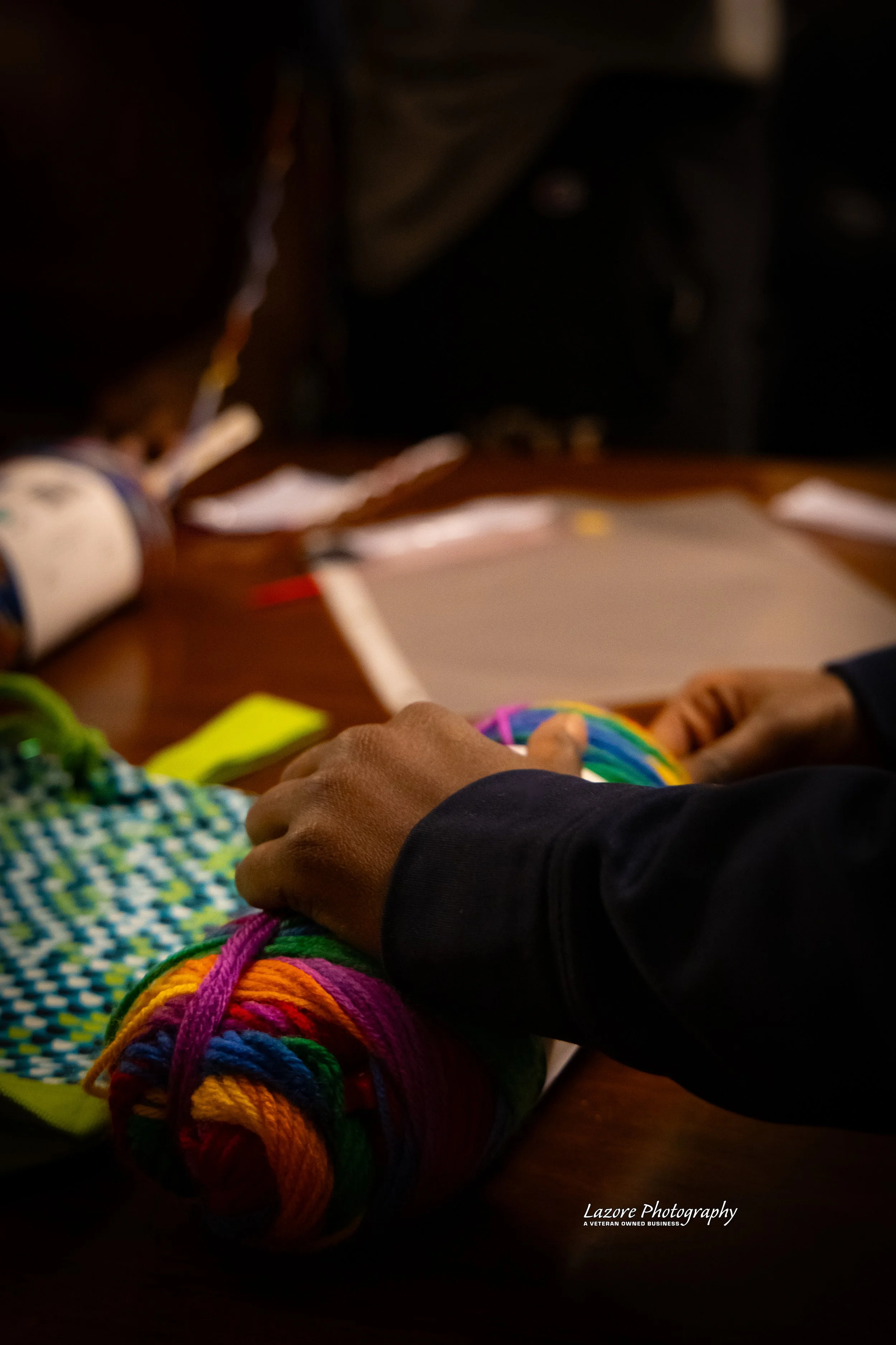 A person holding a multicolored ball of yarn on a wooden table, with other yarn and papers nearby.