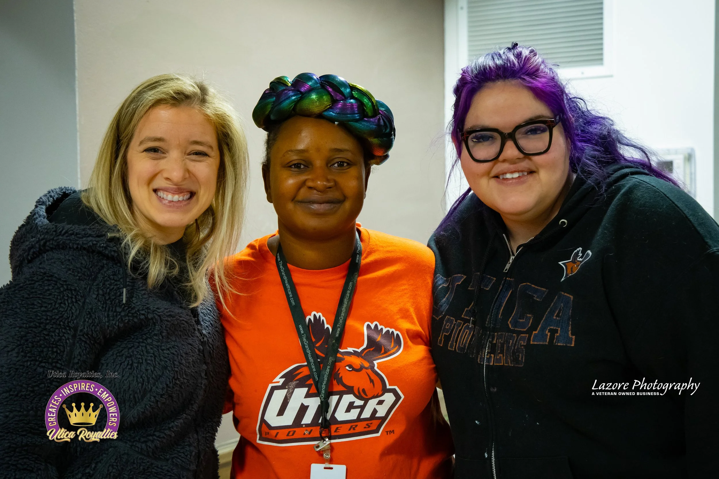 Three women smiling and standing close together indoors. The woman on the left has blonde hair and wears a black fuzzy jacket. The woman in the middle has dark skin, colorful braided hair, and wears an orange T-shirt with a moose logo and text that r