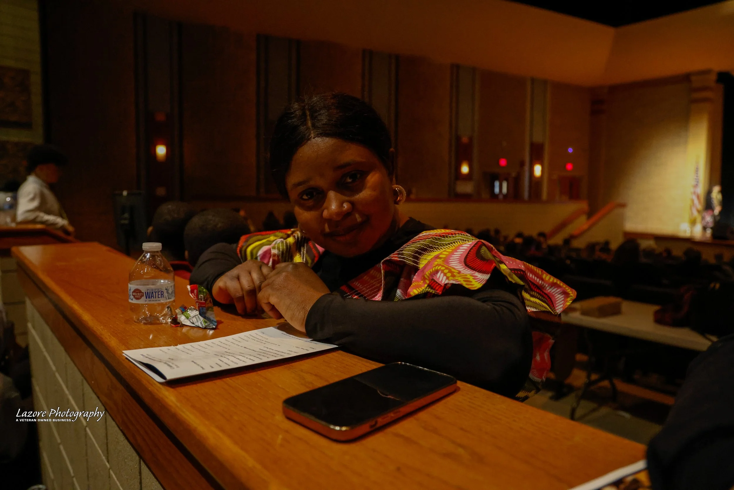 Woman sitting at a wooden counter in an auditorium or conference hall, with a bottle of water, a smartphone, and a sheet of paper in front of her, during an event.