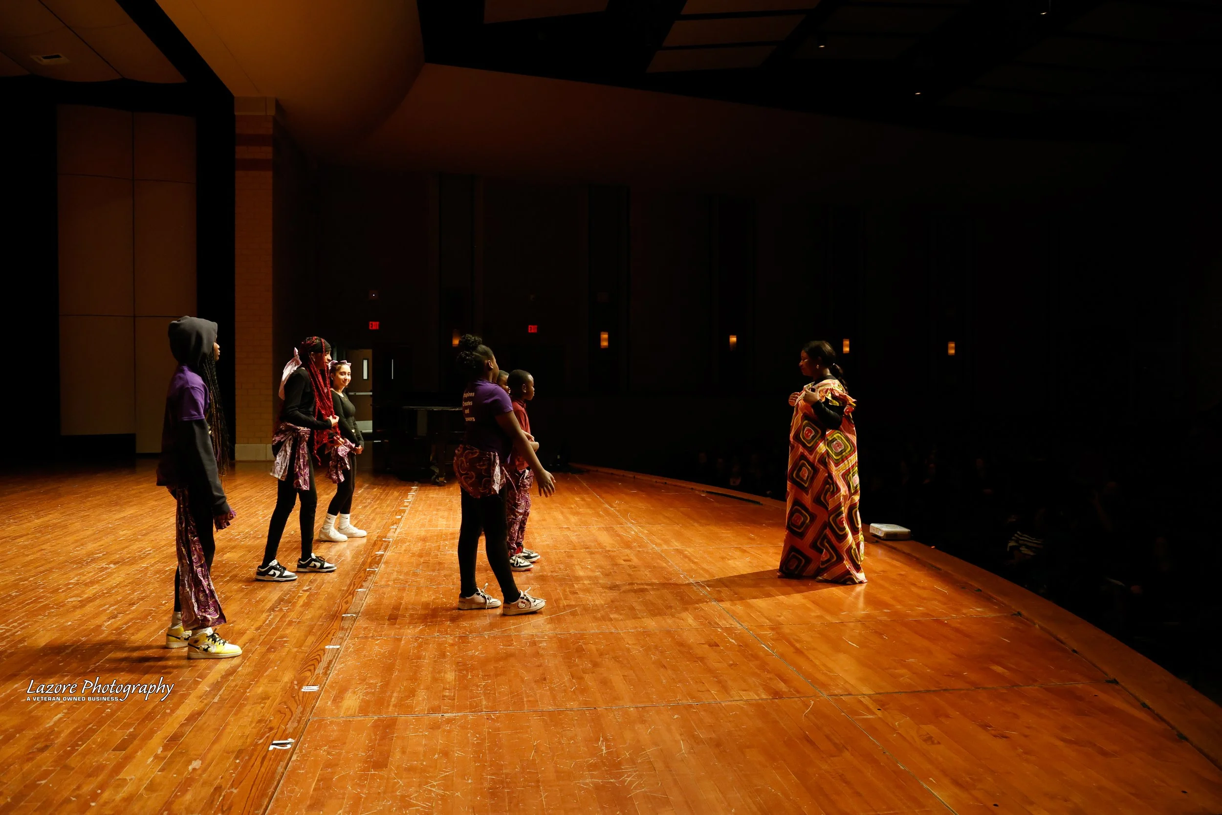 Group of children practicing dance on stage with instructor in a dimly lit auditorium.