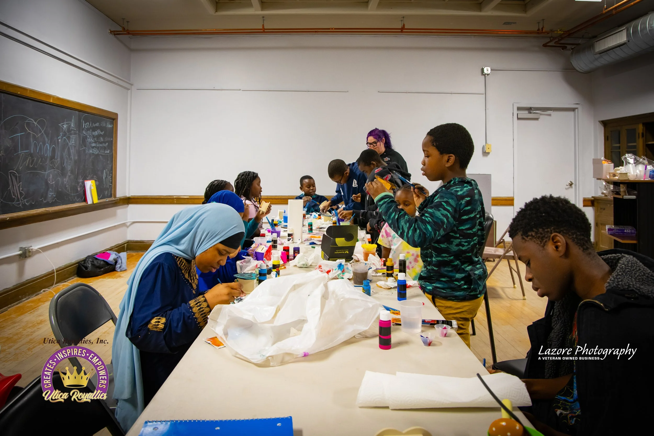 Children and adults gathered around a long table engaging in arts and crafts activities, with various supplies like colored glue bottles and paper, in a classroom or community center setting.