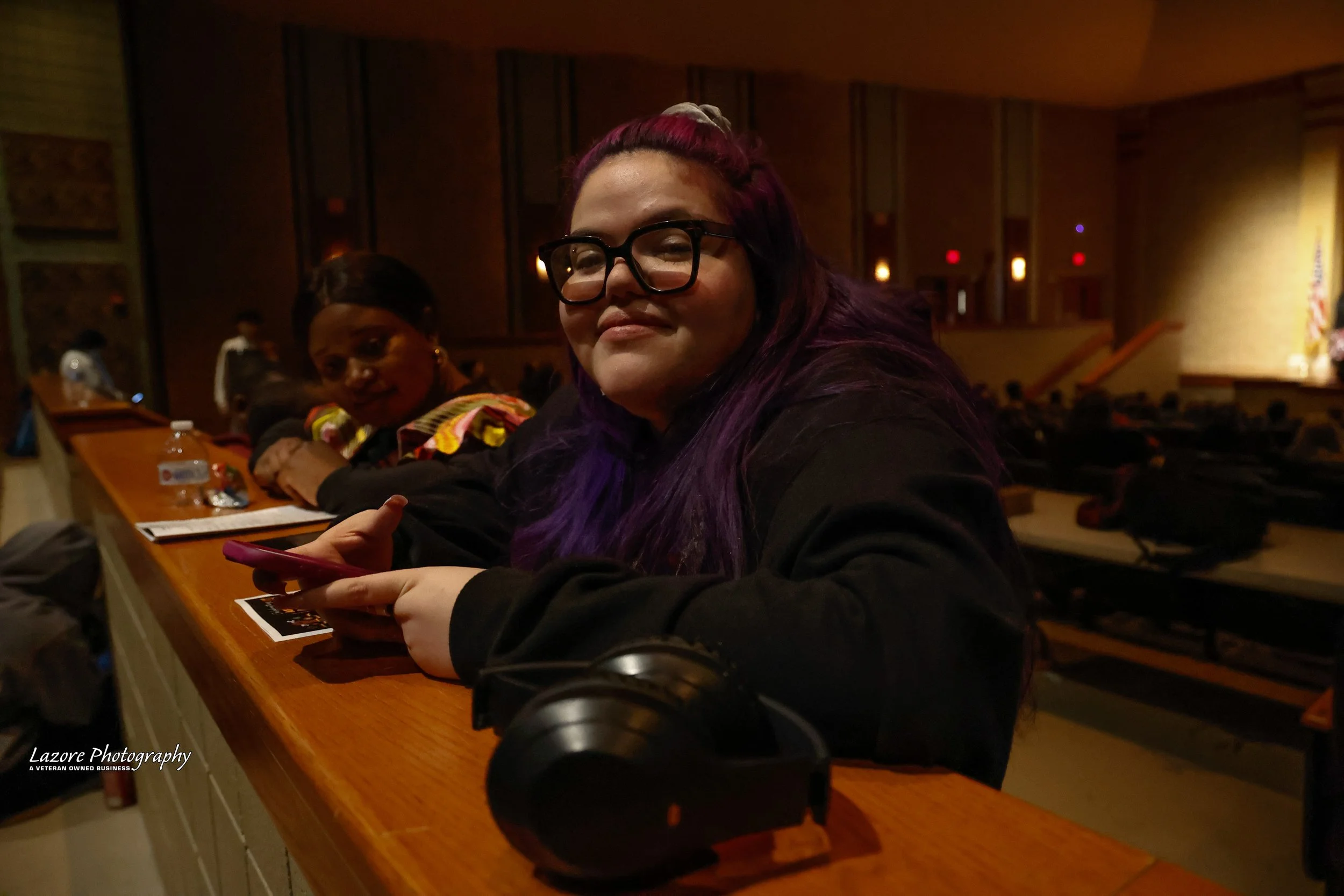 Woman with purple hair and glasses sitting at a wooden table in a dimly lit auditorium, looking at the camera, with another woman behind her. Items on the table include a phone, headphones, and a water bottle.