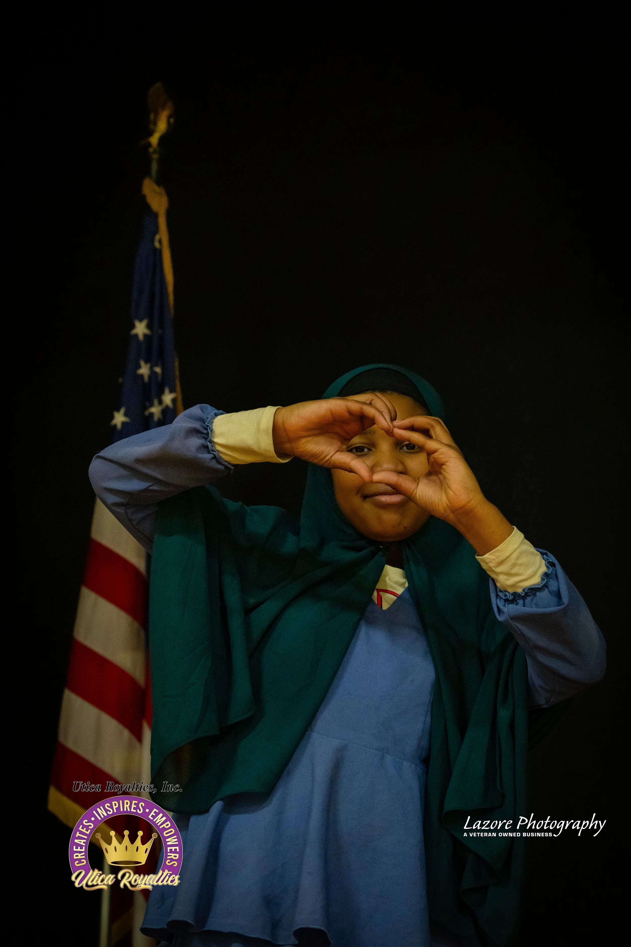 Young girl wearing a blue and yellow school uniform and a headscarf, making a heart shape with her hands around her eye, standing in front of an American flag.