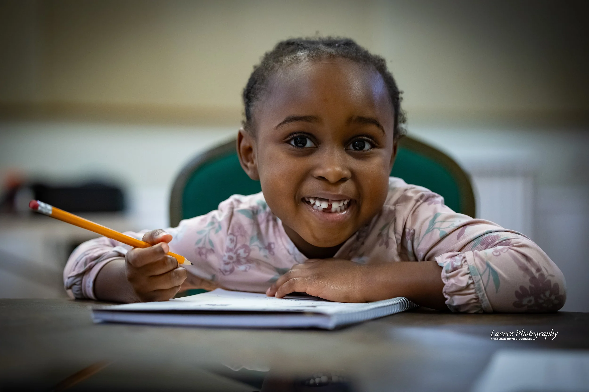 A young girl smiling while sitting at a table with a notebook and pencil, in a classroom setting.