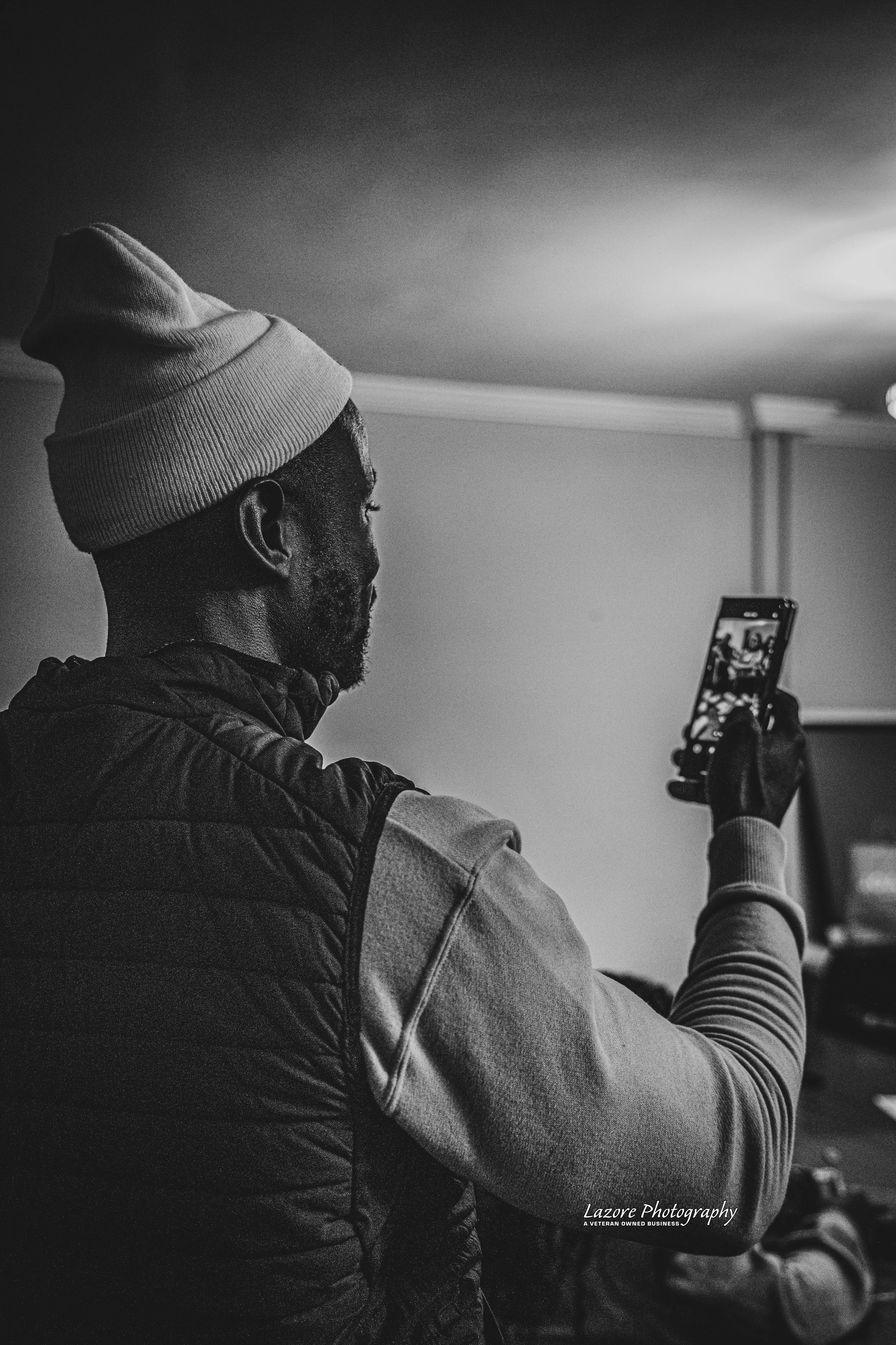 A man with a beanie and a puffy vest taking a selfie with a smartphone in a dimly lit room.