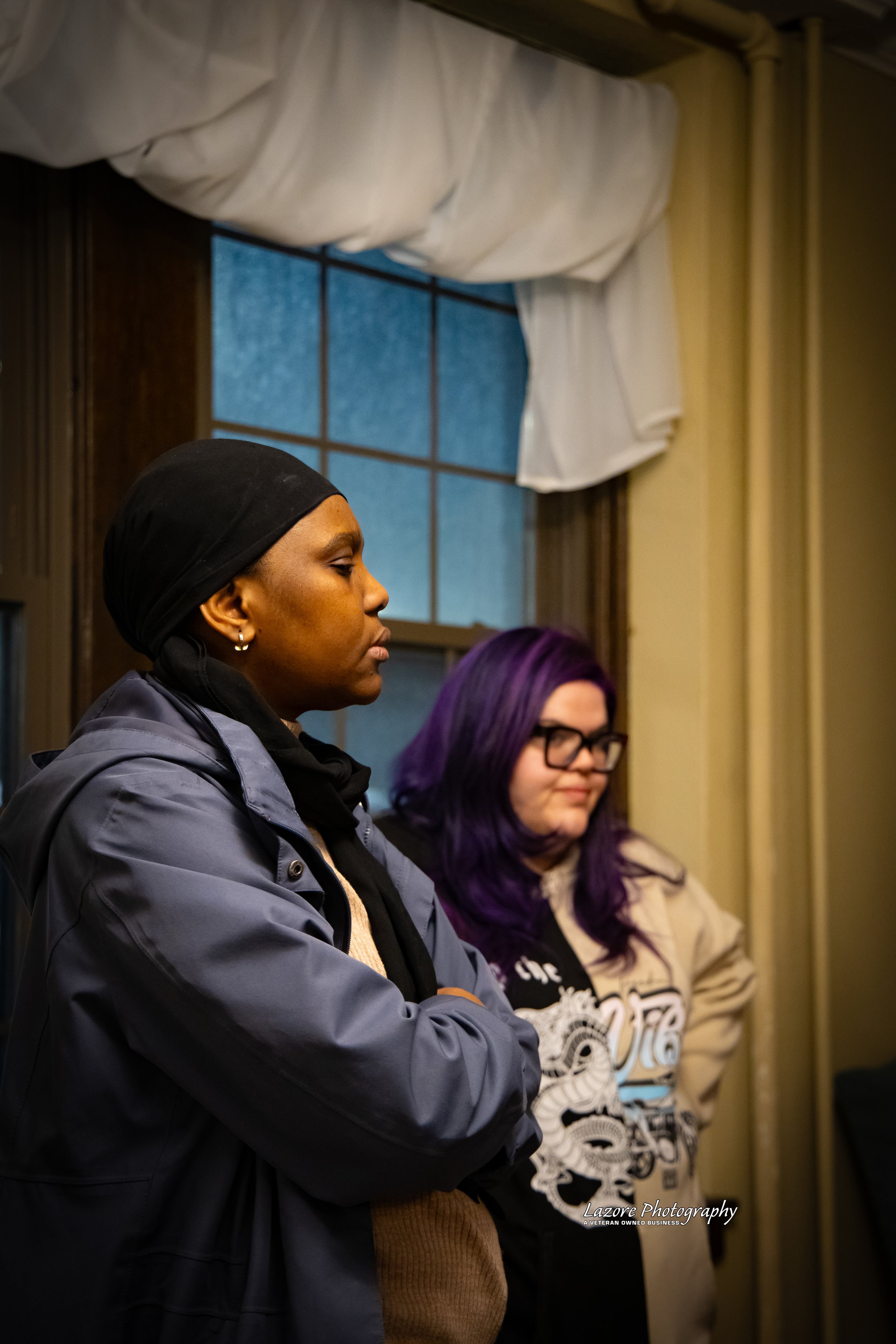 Two women standing indoors near a window, one with a black headscarf and glasses, and the other with purple hair and glasses, both appearing focused or contemplative.
