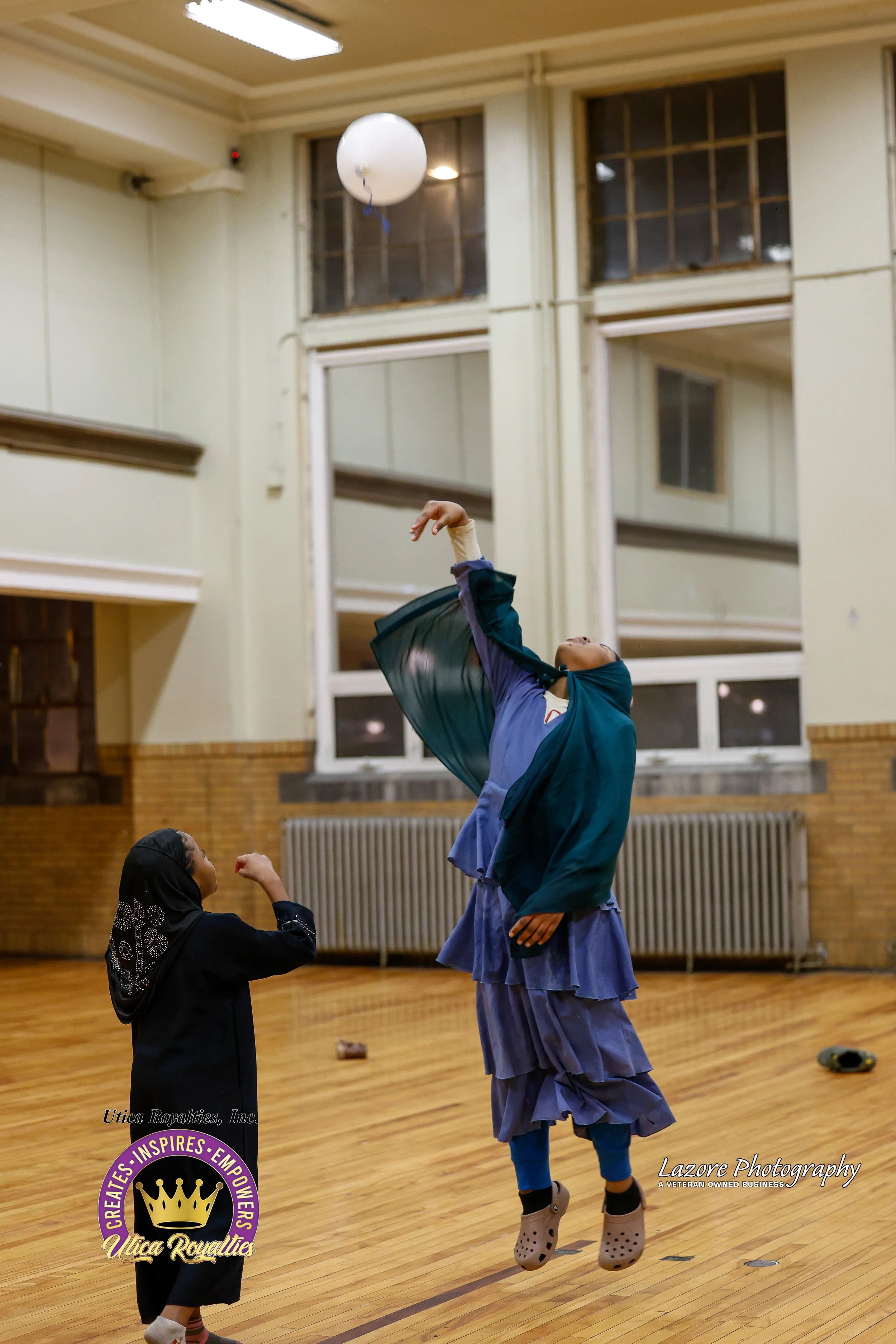Two girls, one in hijab and one in a blue dress, playing with a balloon in a gymnasium with wooden floors and large windows.