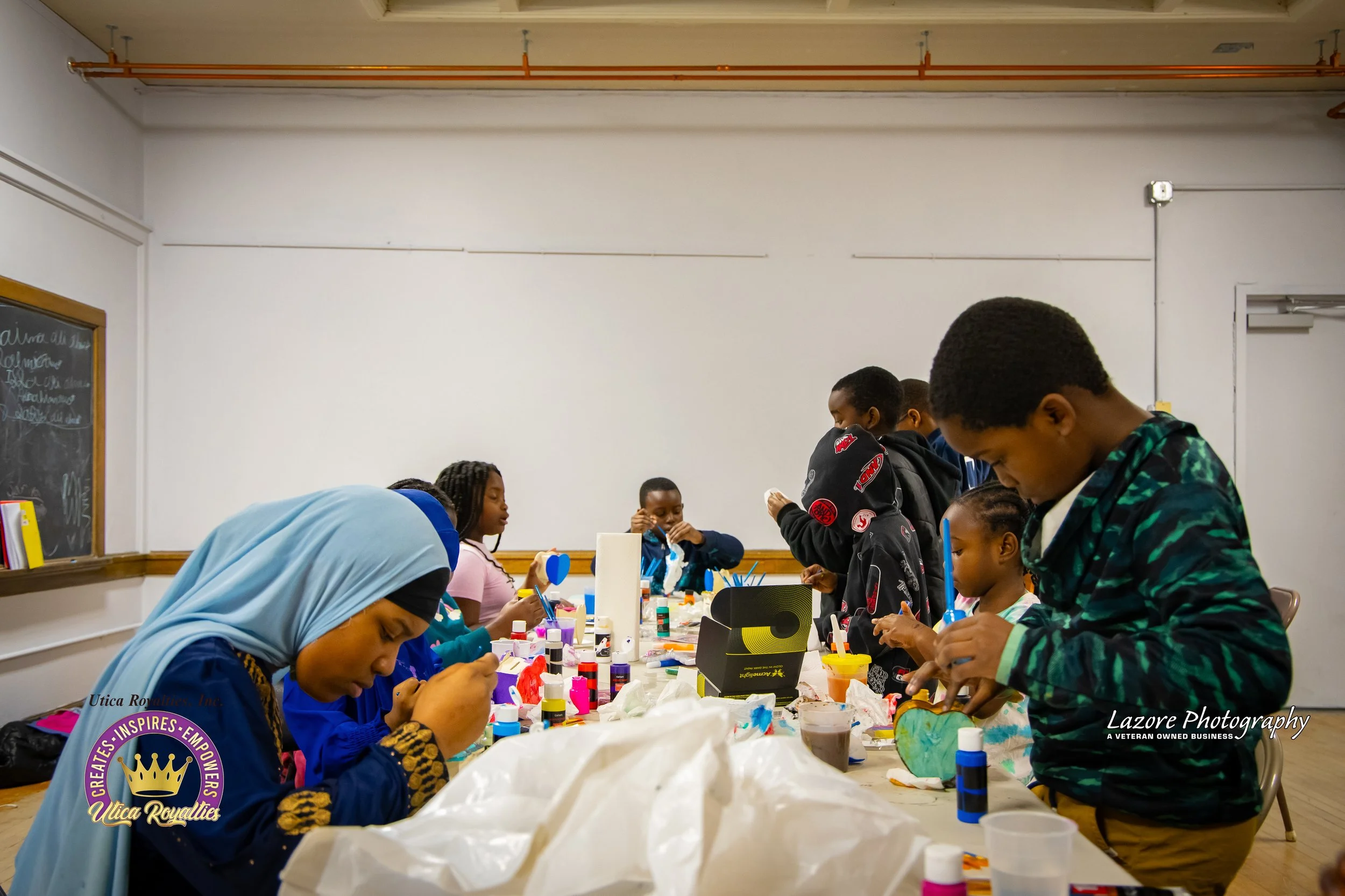 Children and teenagers engaged in arts and crafts at a long table, working with paints, glue, and paper in a classroom setting.