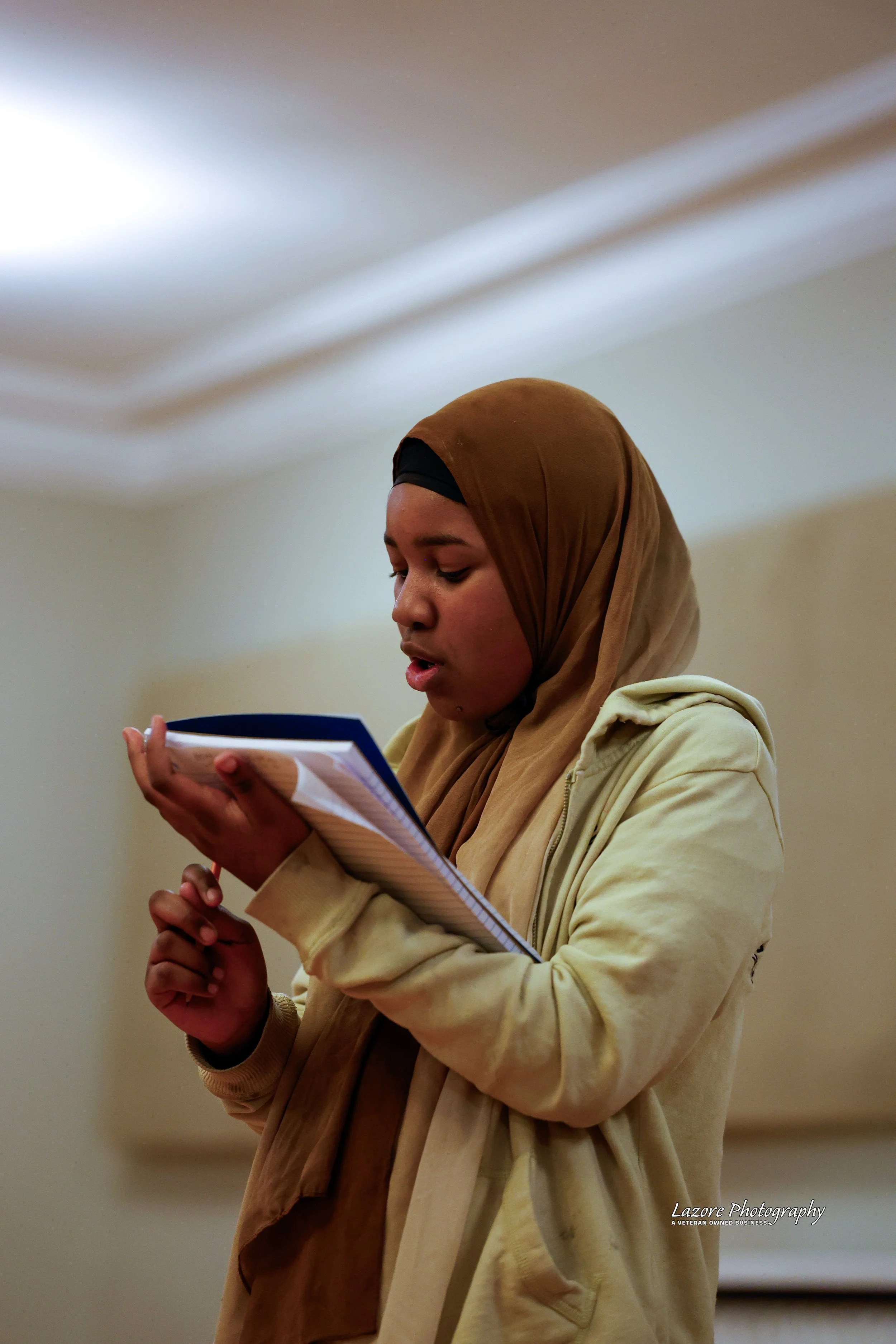 A woman wearing a beige jacket and a brown headscarf reading from a notebook.