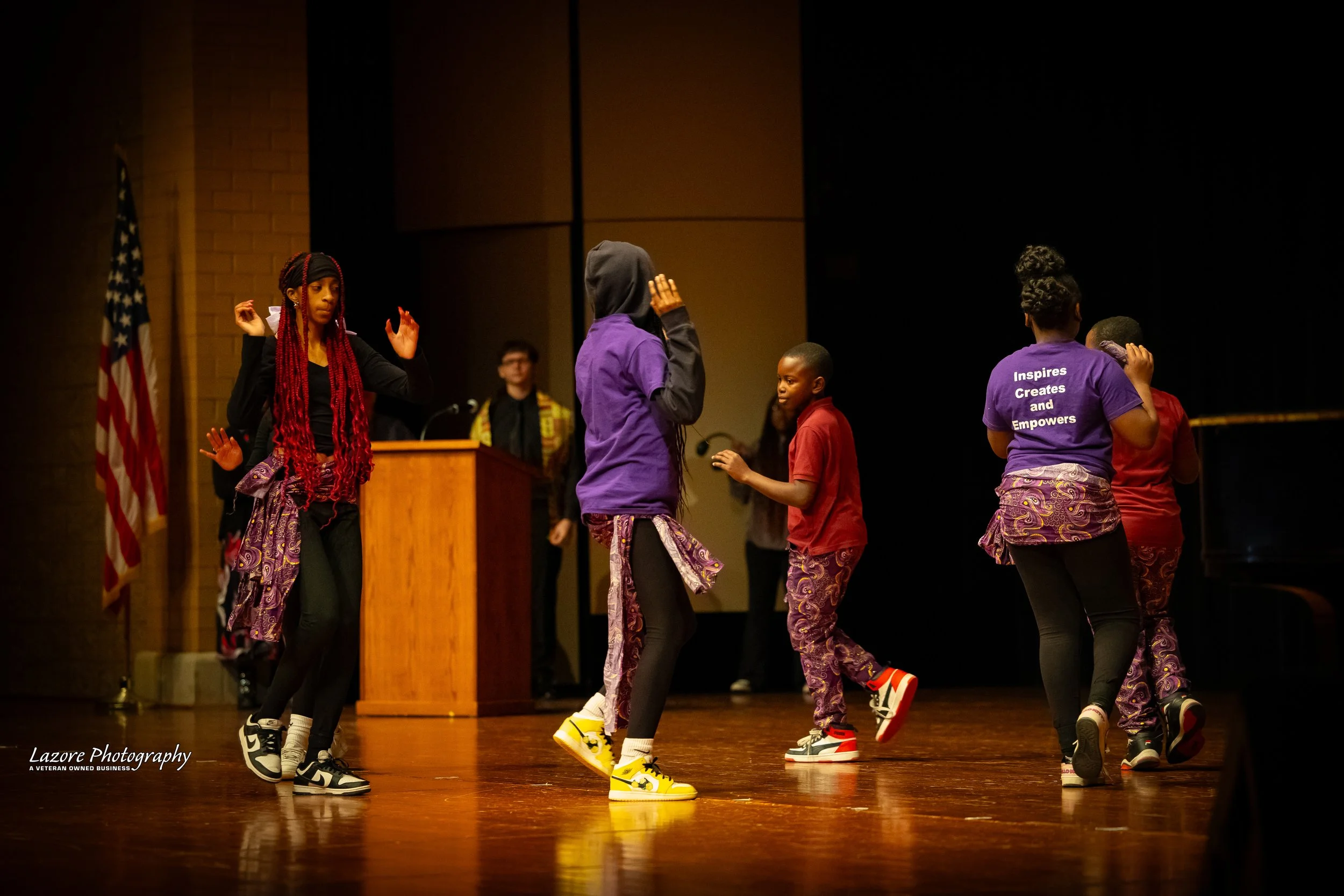 Group of children dancing on stage with a woman in a purple shirt and an American flag in the background.