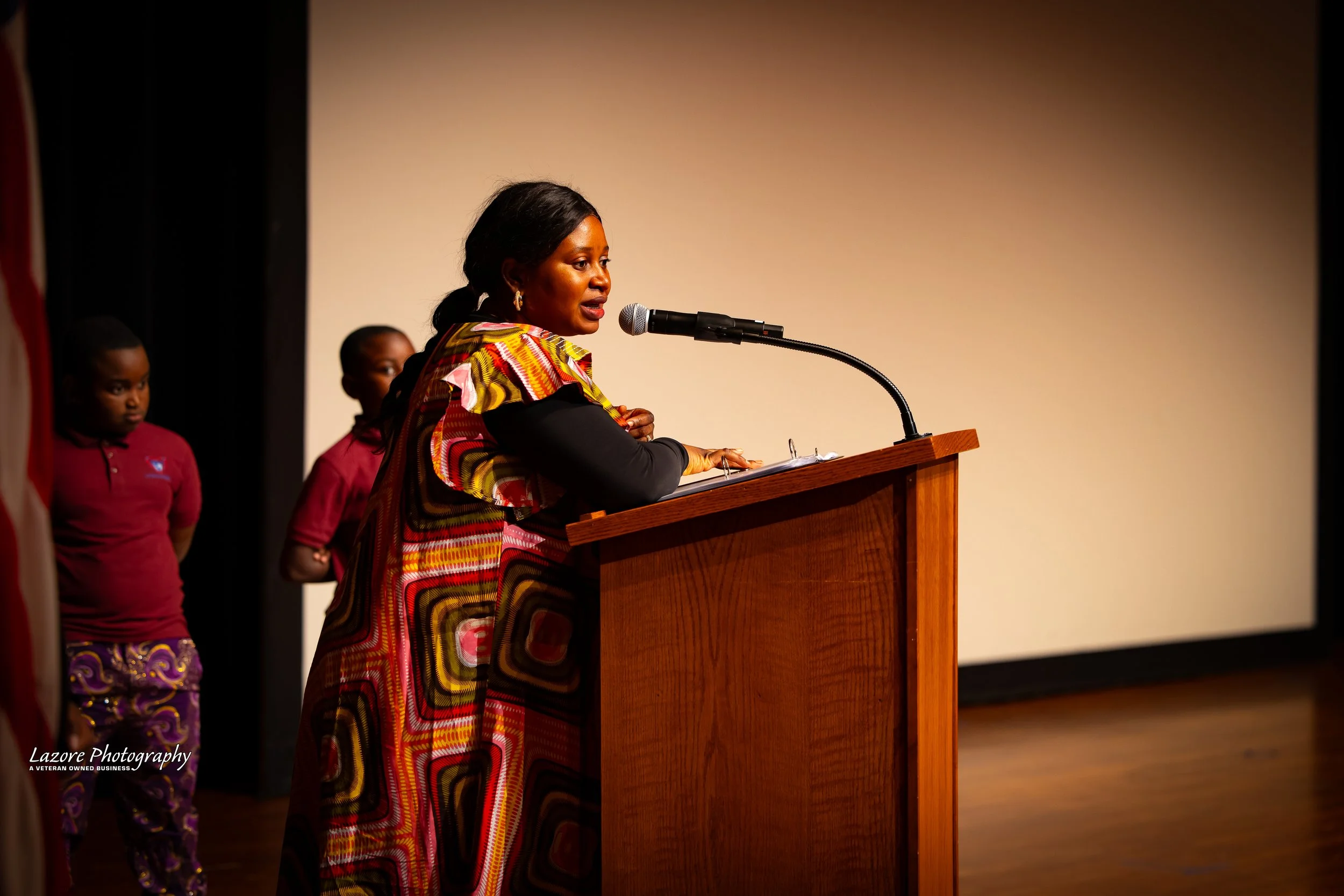 A woman speaking at a podium with a microphone, wearing a colorful patterned dress, with two children standing behind her on stage.