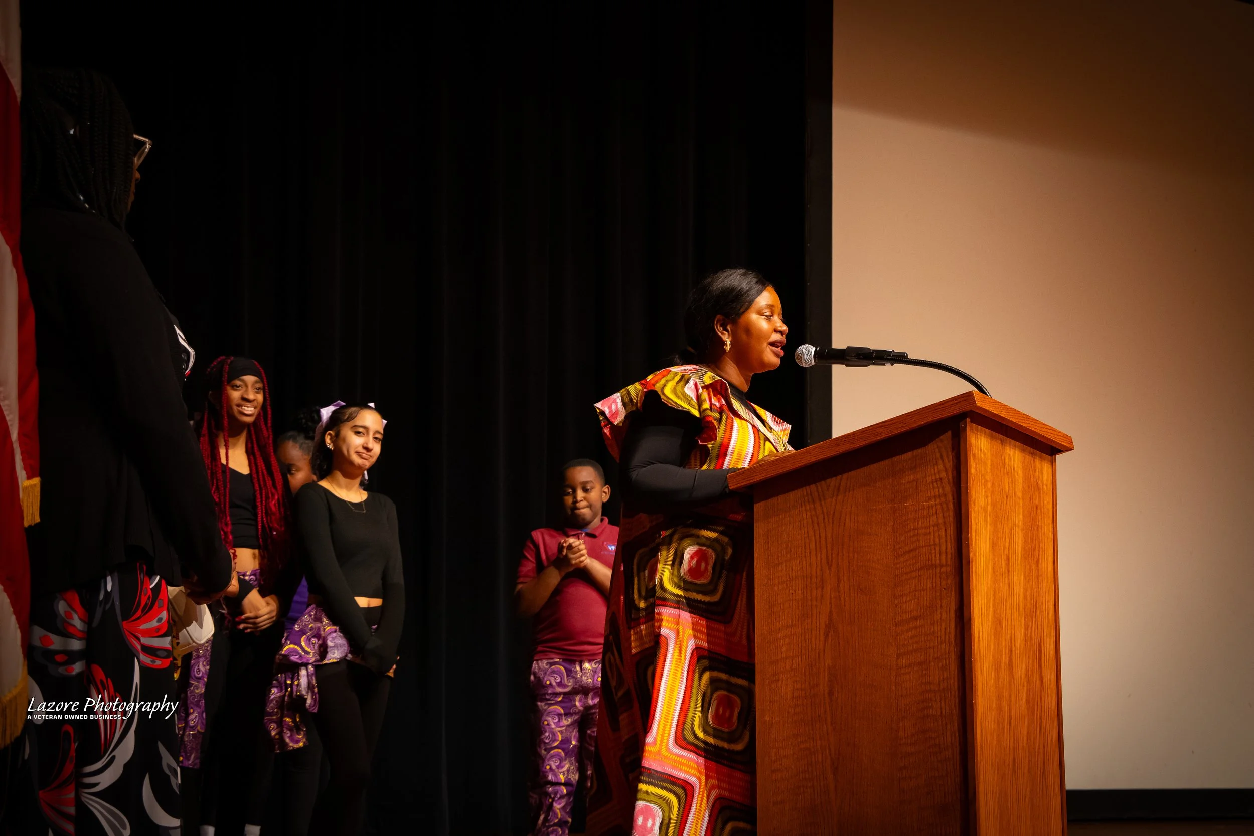 Woman speaking at a wooden podium with children standing in a line behind her on a stage with black curtains.