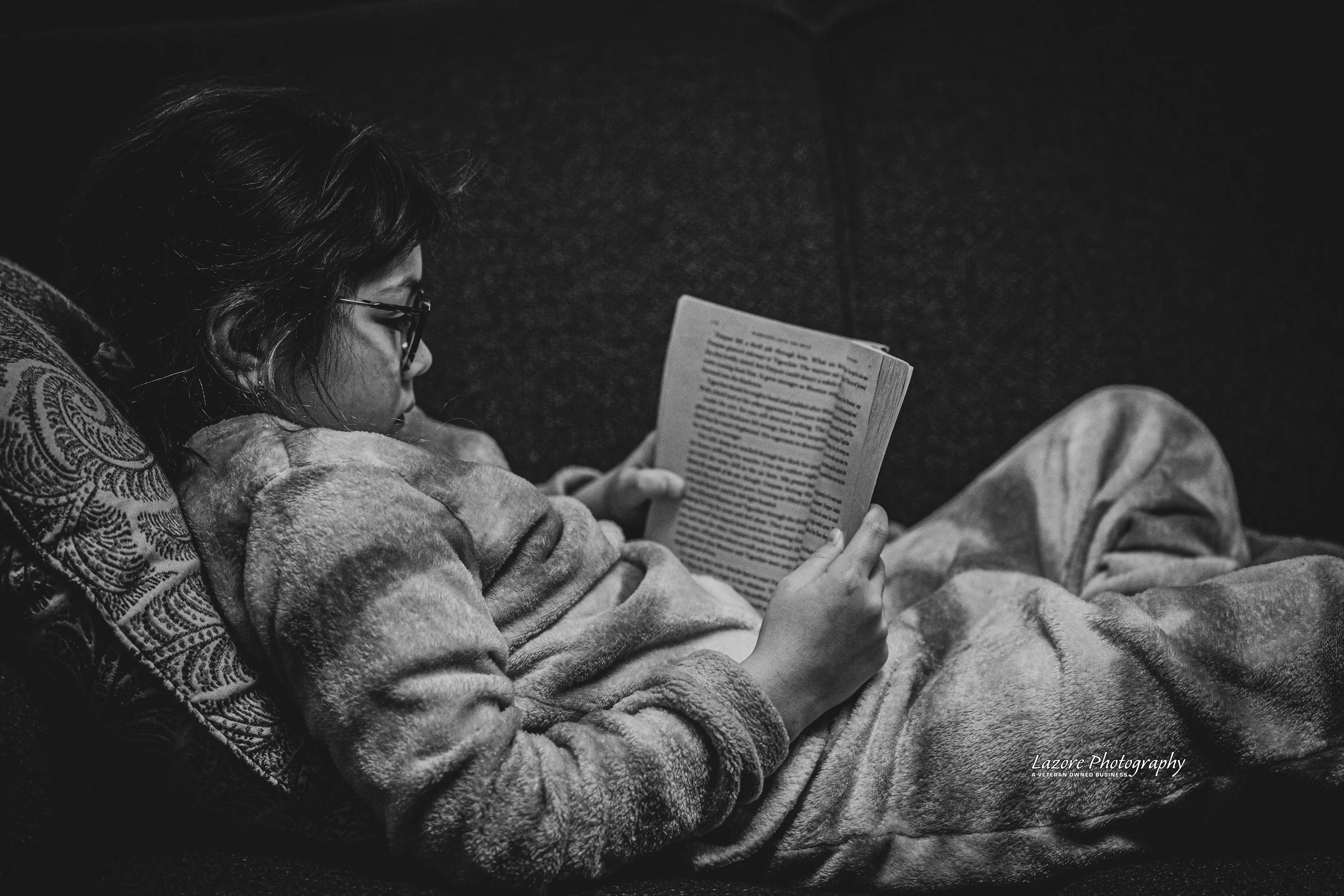 A young girl with glasses and a patterned hoodie reading a book while lying on a couch with a blanket, in black and white.