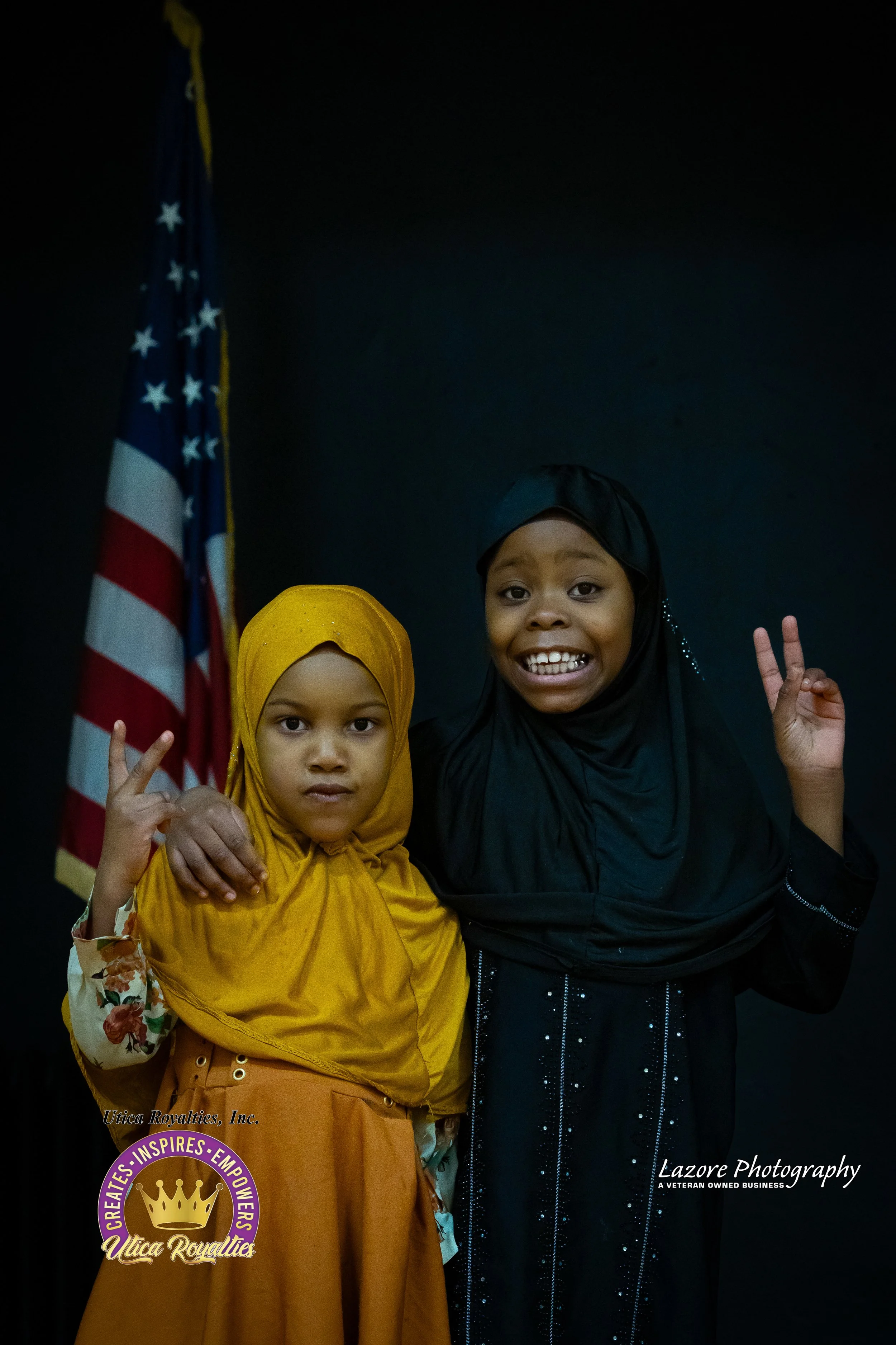 Two smiling young girls, one in a yellow headscarf and the other in a black headscarf, posing together with one making a peace sign, standing in front of an American flag.