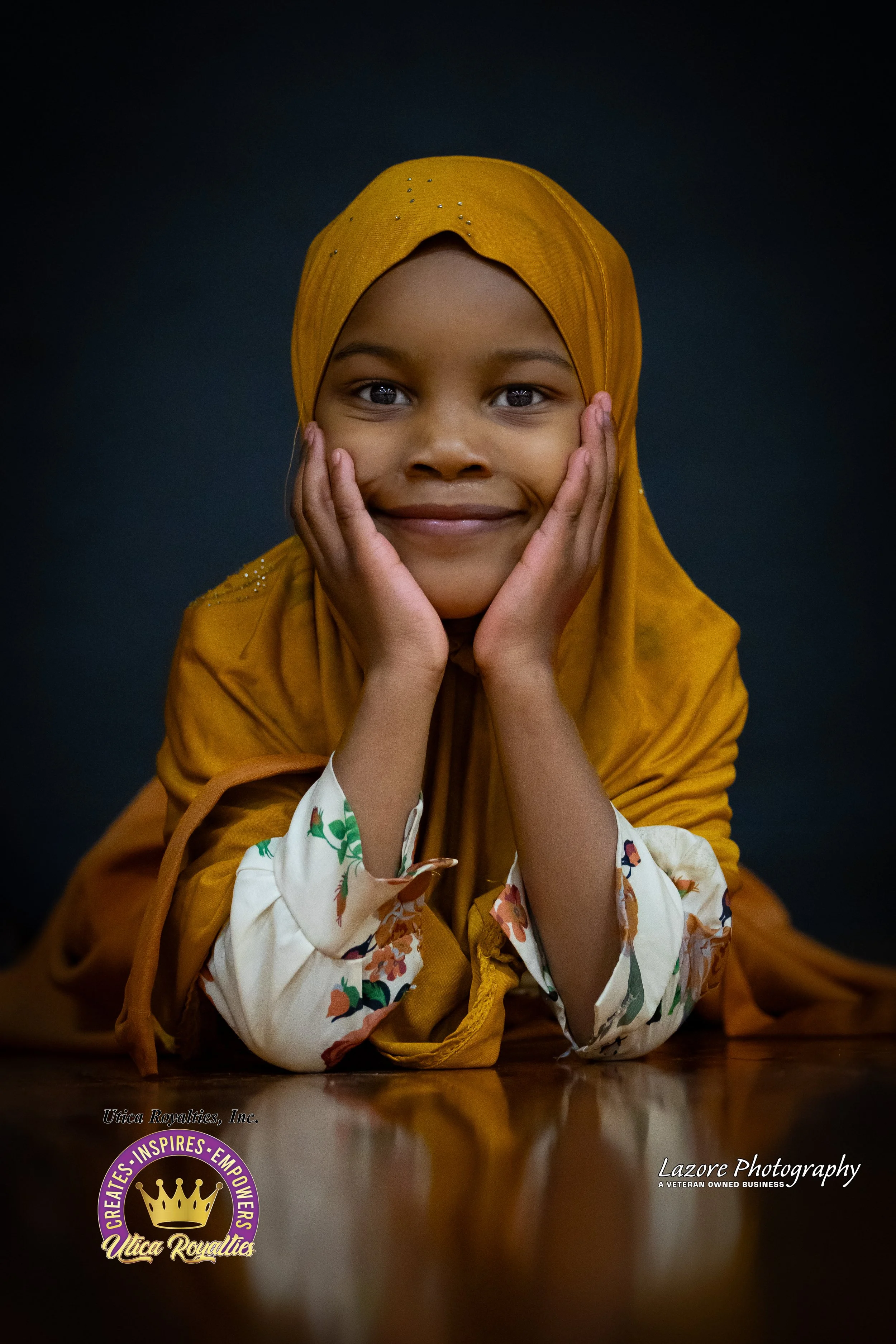 A young girl wearing a yellow hijab with small embellishments, smiling and resting her face in her hands, against a dark background.