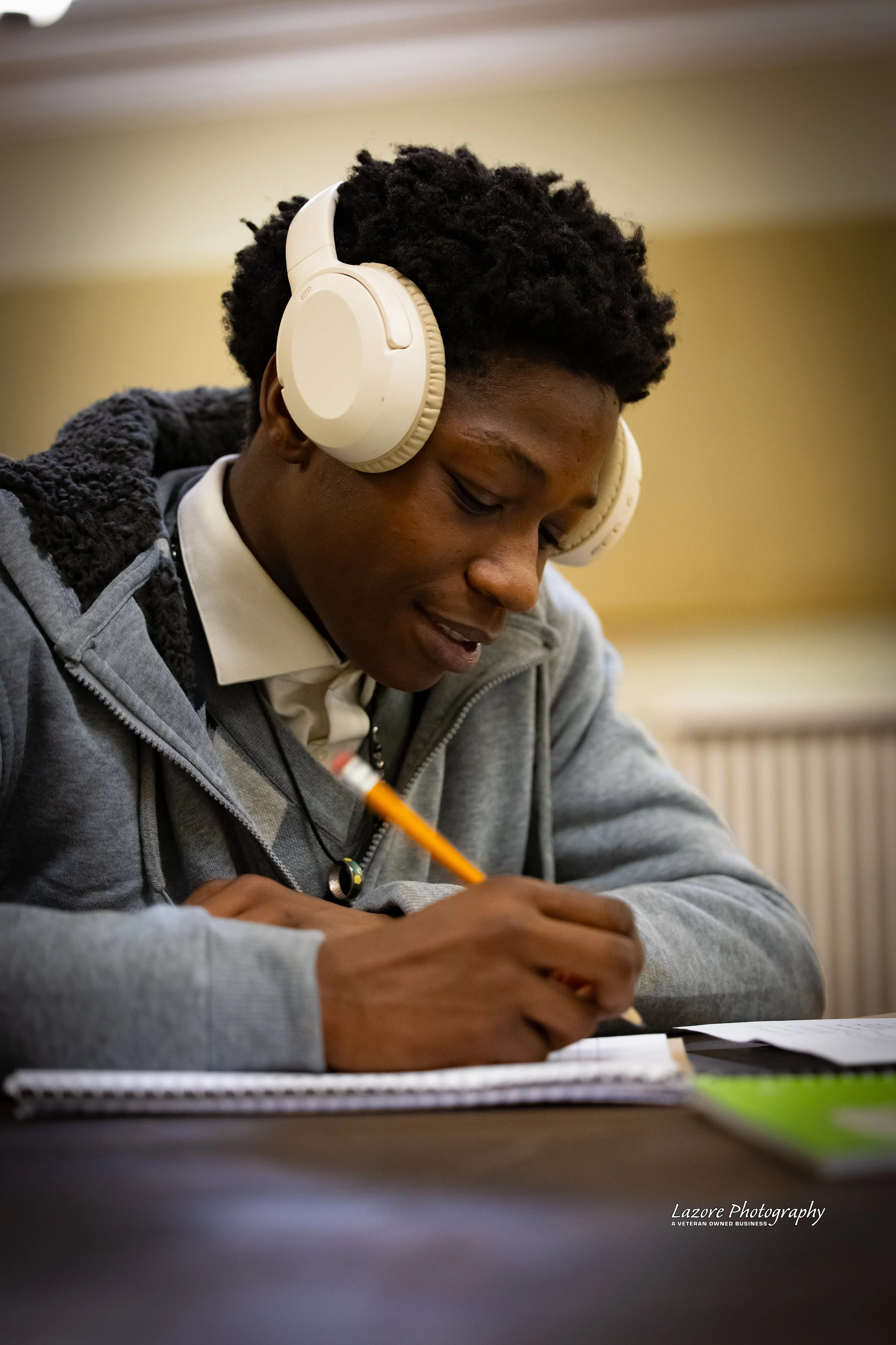 A young man with curly hair wearing large cream-colored headphones, writing in a spiral notebook with a yellow pencil, seated at a desk indoors.