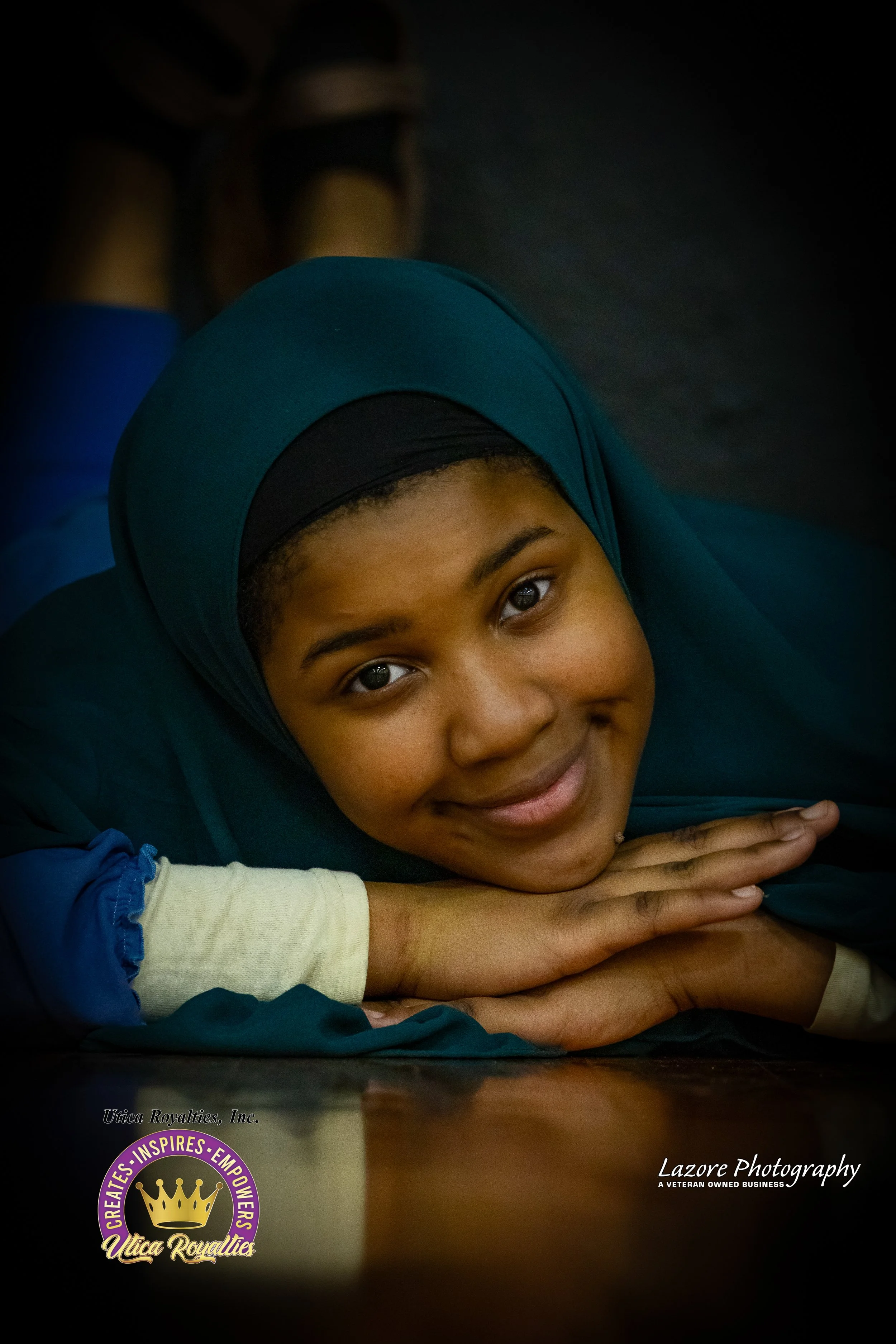 Young girl in a teal hijab lying on a wooden surface, smiling at the camera.