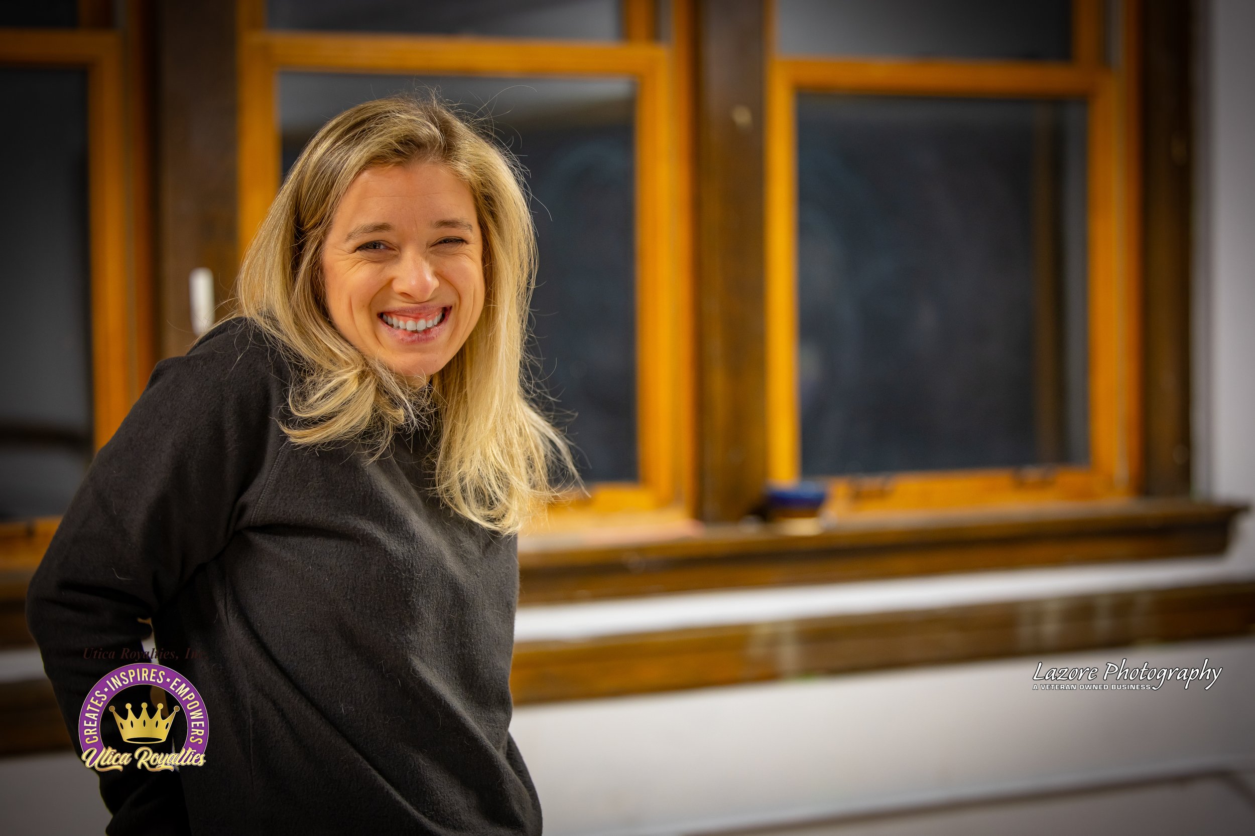 A smiling woman with blonde hair wearing a black sweater, standing indoors near a window with wooden frames, with a piano in the background.