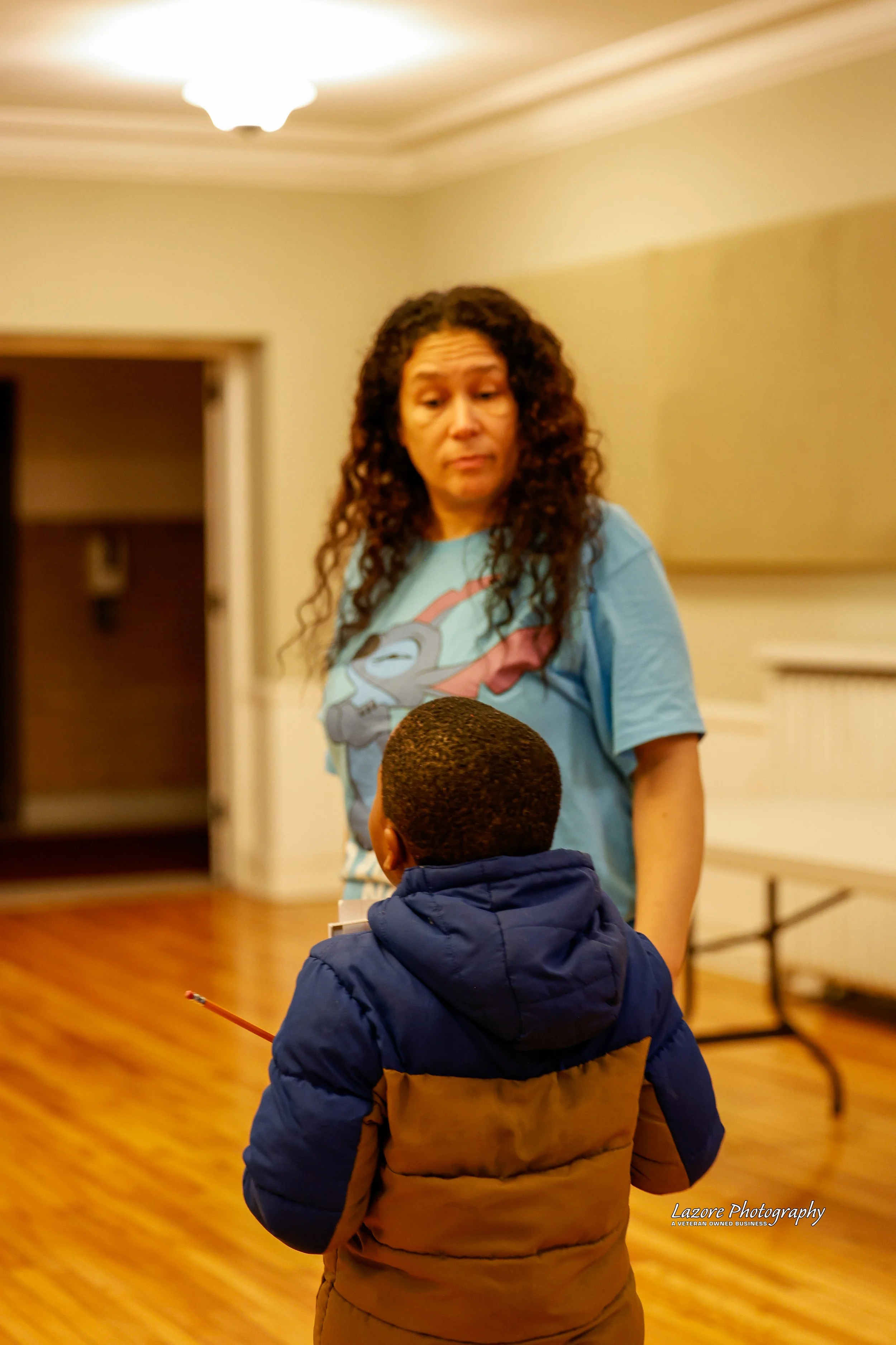 A woman with long curly hair looking at a young boy with short hair, dressed in a blue and tan winter jacket, in an indoor setting with wood floor and light-colored walls.